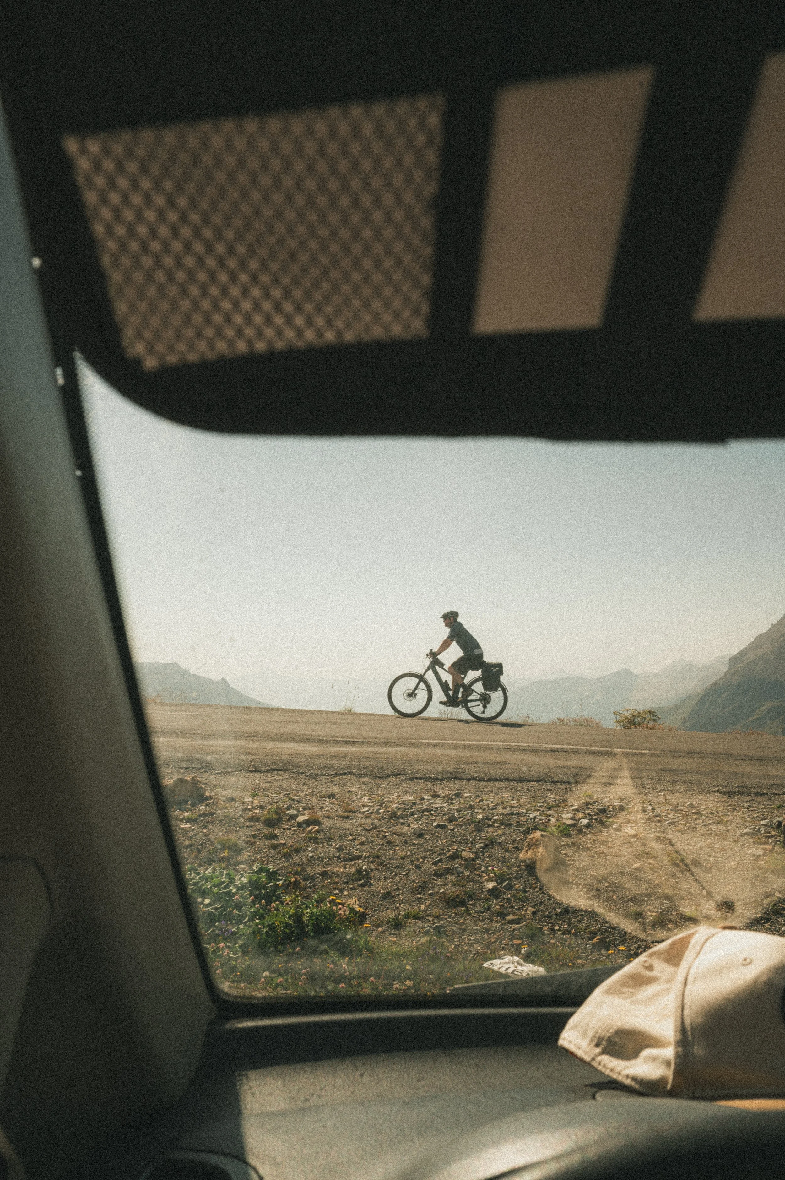 Vue à travers le pare-brise d'une voiture montrant un cycliste pédalant sur une route de campagne avec un paysage de montagnes en arrière-plan.