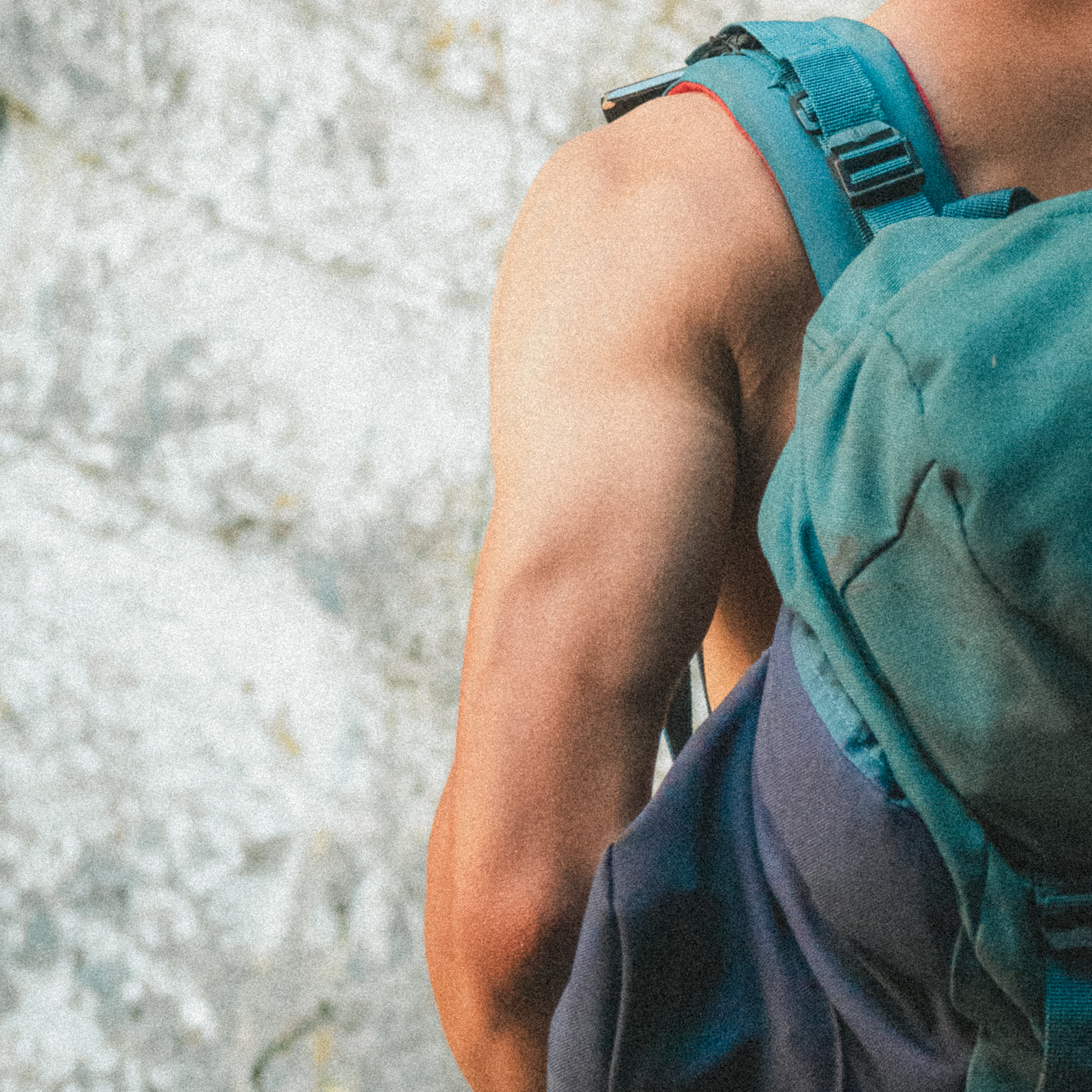 Partie du haut du corps d'une personne portant un sac à dos bleu avec un bracelet bleu, en position penchée vers l'avant, contre un fond de surface rocheuse ou en béton.