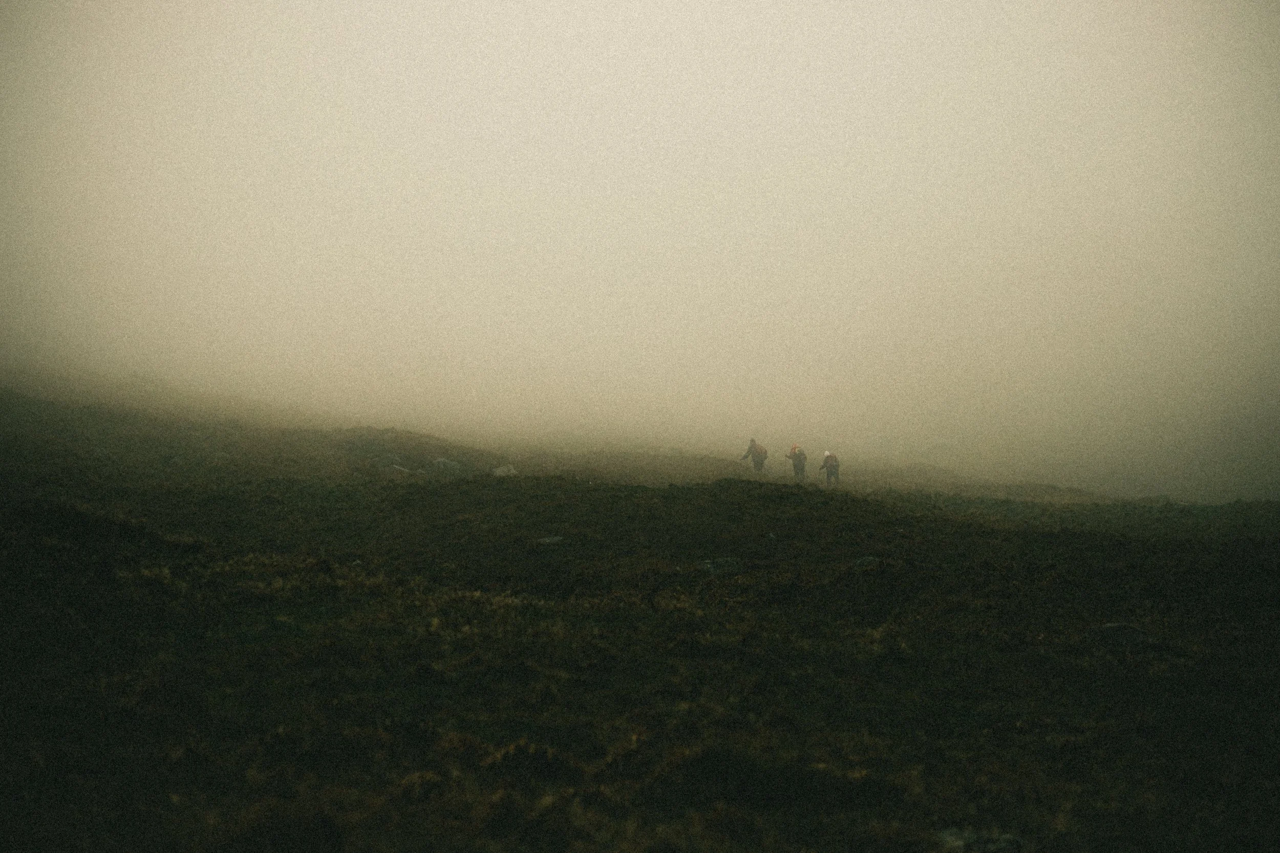 Trois randonneurs marchant dans une zone brumeuse et sombre, avec un ciel gris. Le paysage est rocheux et dépourvu de végétation visible, évoquant un décor de haute montagne ou de terrain volcanique.