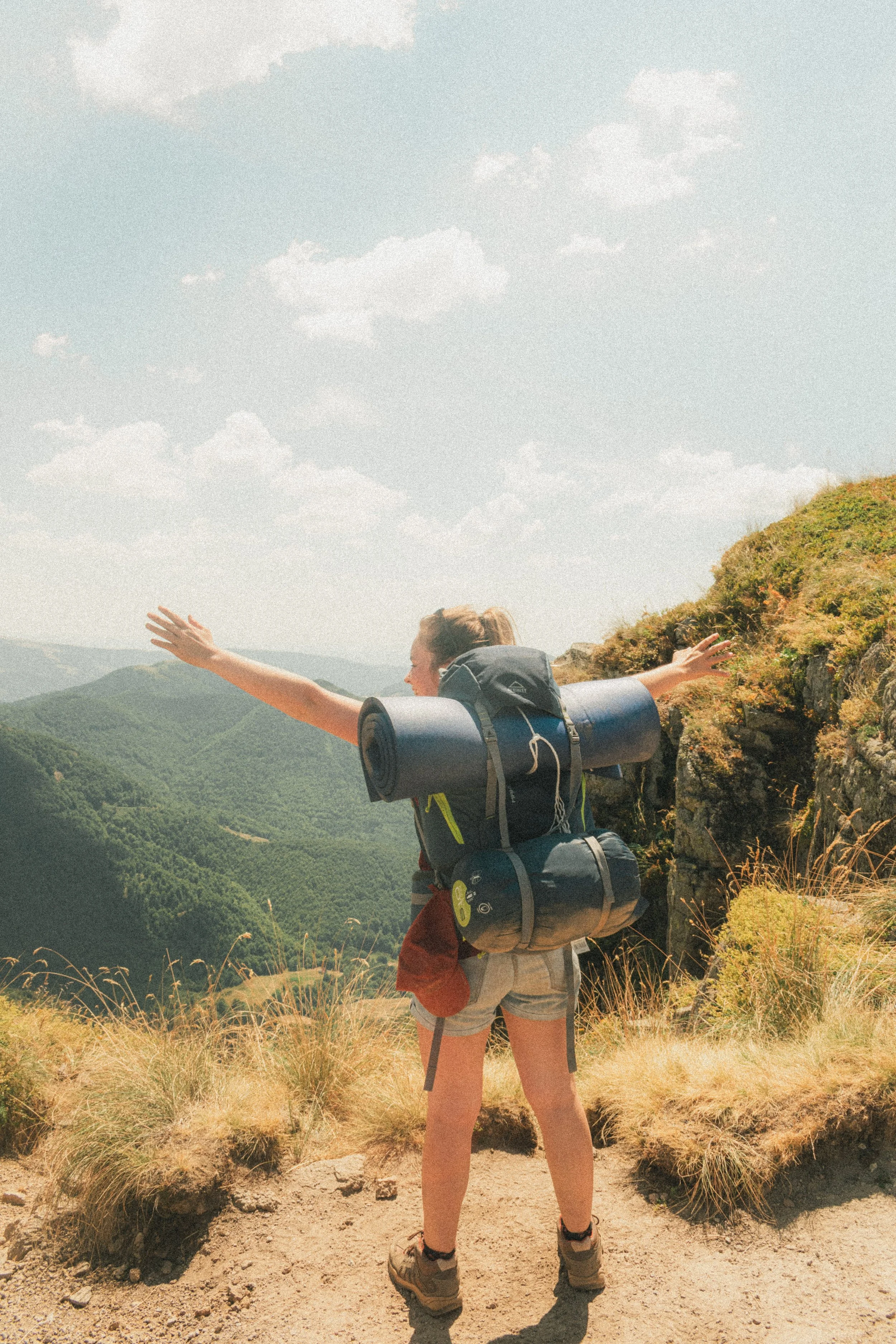Une femme randonneuse avec un sac à dos et un tapis de sol profite d'une vue panoramique depuis une montagne, levant les bras avec un paysage de montagnes verdoyantes en arrière-plan.
