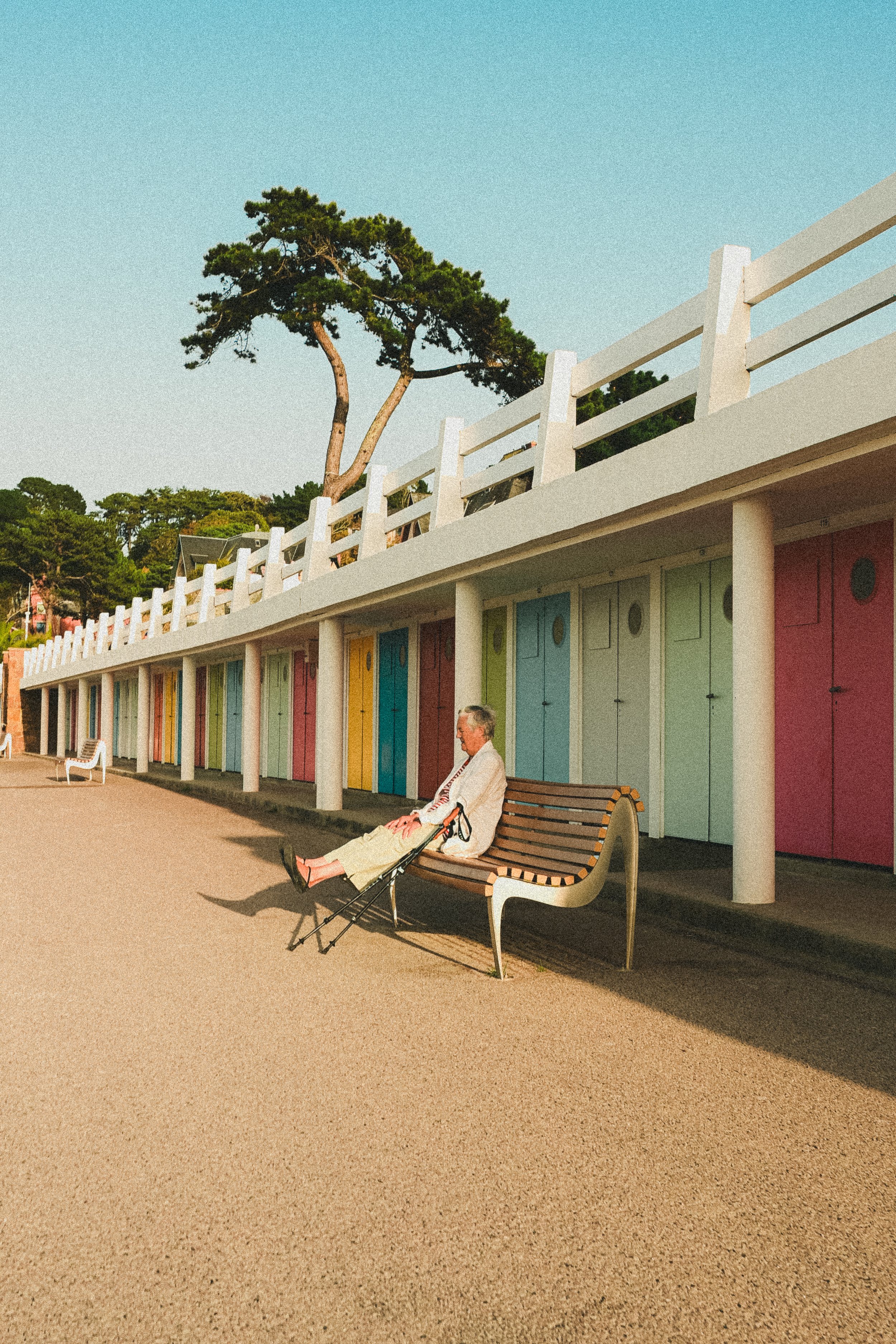 Une personne assise sur un banc en bois sous un bâtiment coloré avec des cabines de plage peinte en différentes couleurs, un arbre en arrière-plan, un ciel clair, lors d'une journée ensoleillée.