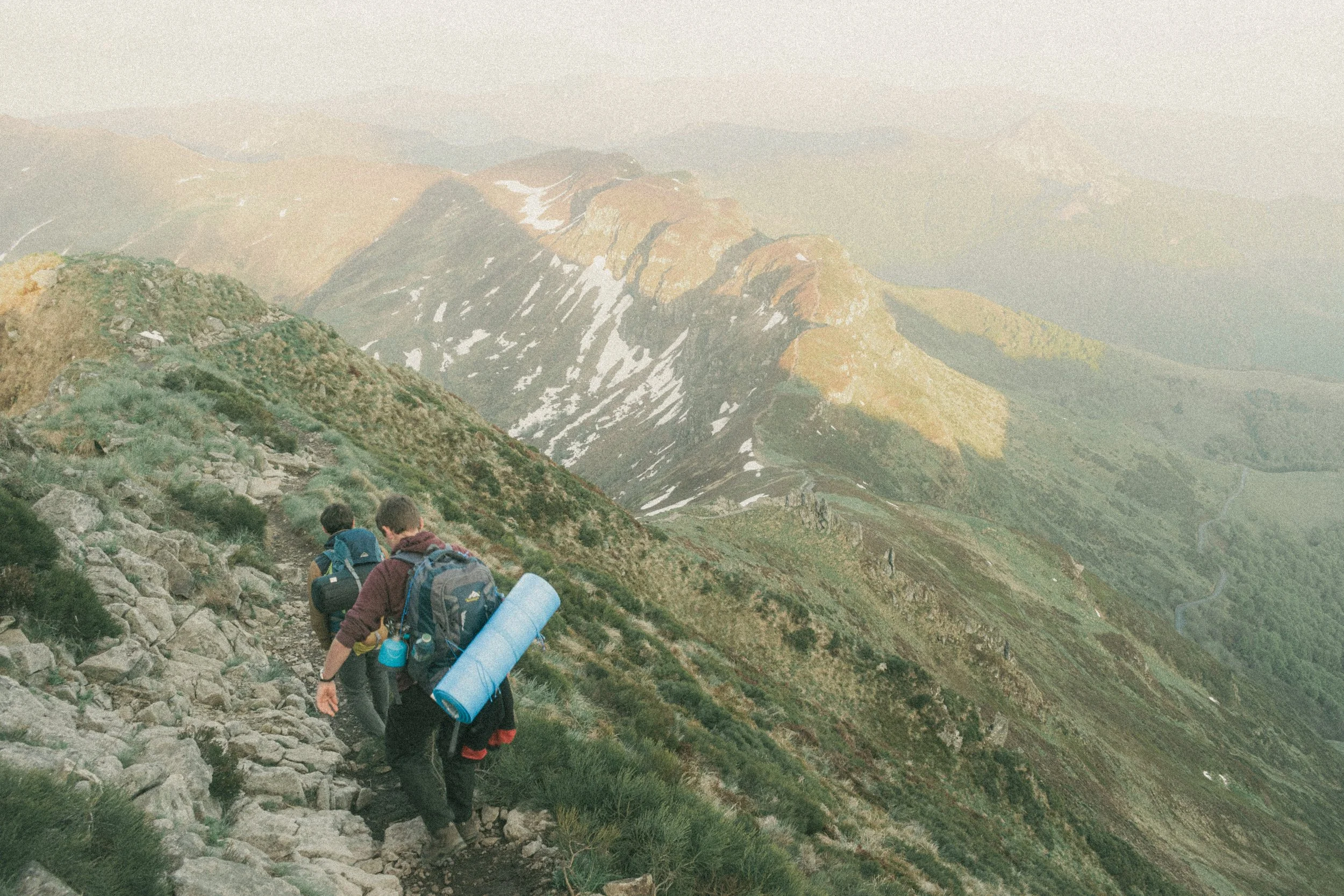 Trois randonneurs marchant sur un sentier de montagne escarpé, avec des sacs à dos, dans un paysage de montagnes verdoyantes avec des taches de neige en arrière-plan.