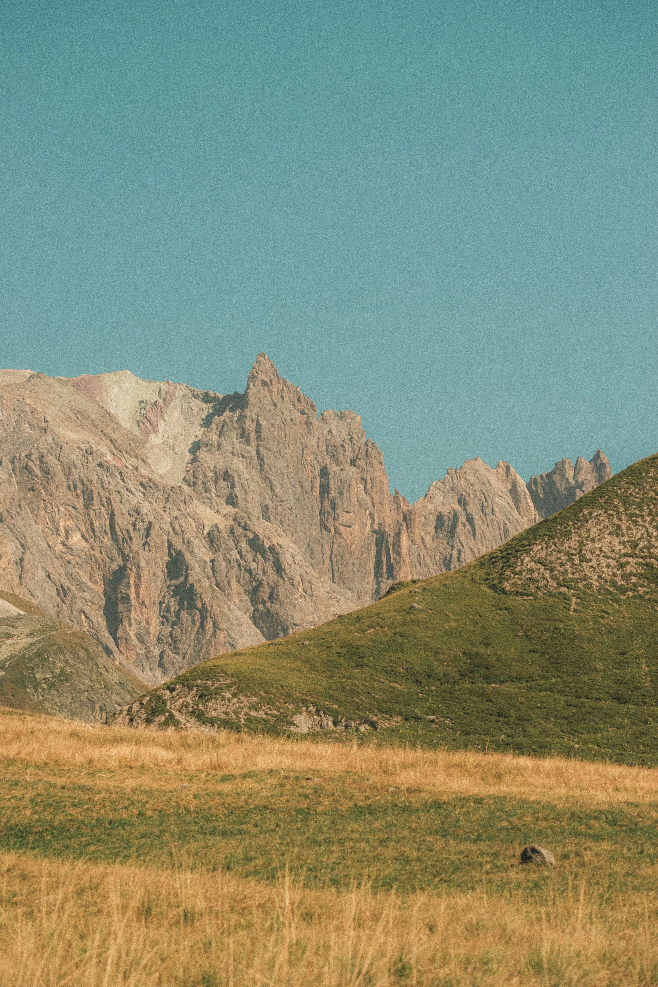Paysage de montagnes avec un ciel bleu clair, des sommets rocheux et une prairie verte au premier plan.