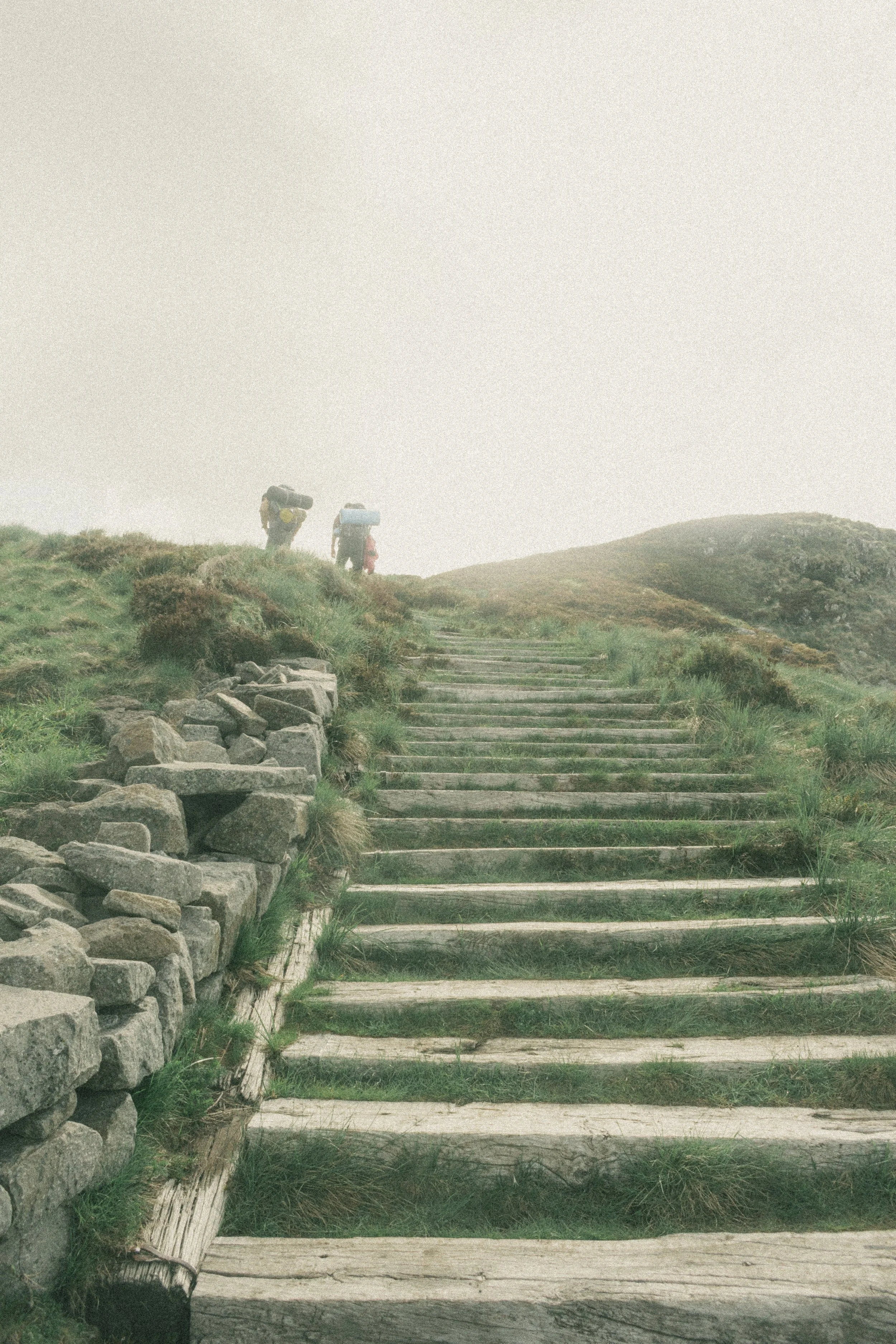 Deux randonneurs portant des sacs à dos gravissent un sentier en escaliers dans une région montagneuse brumeuse.