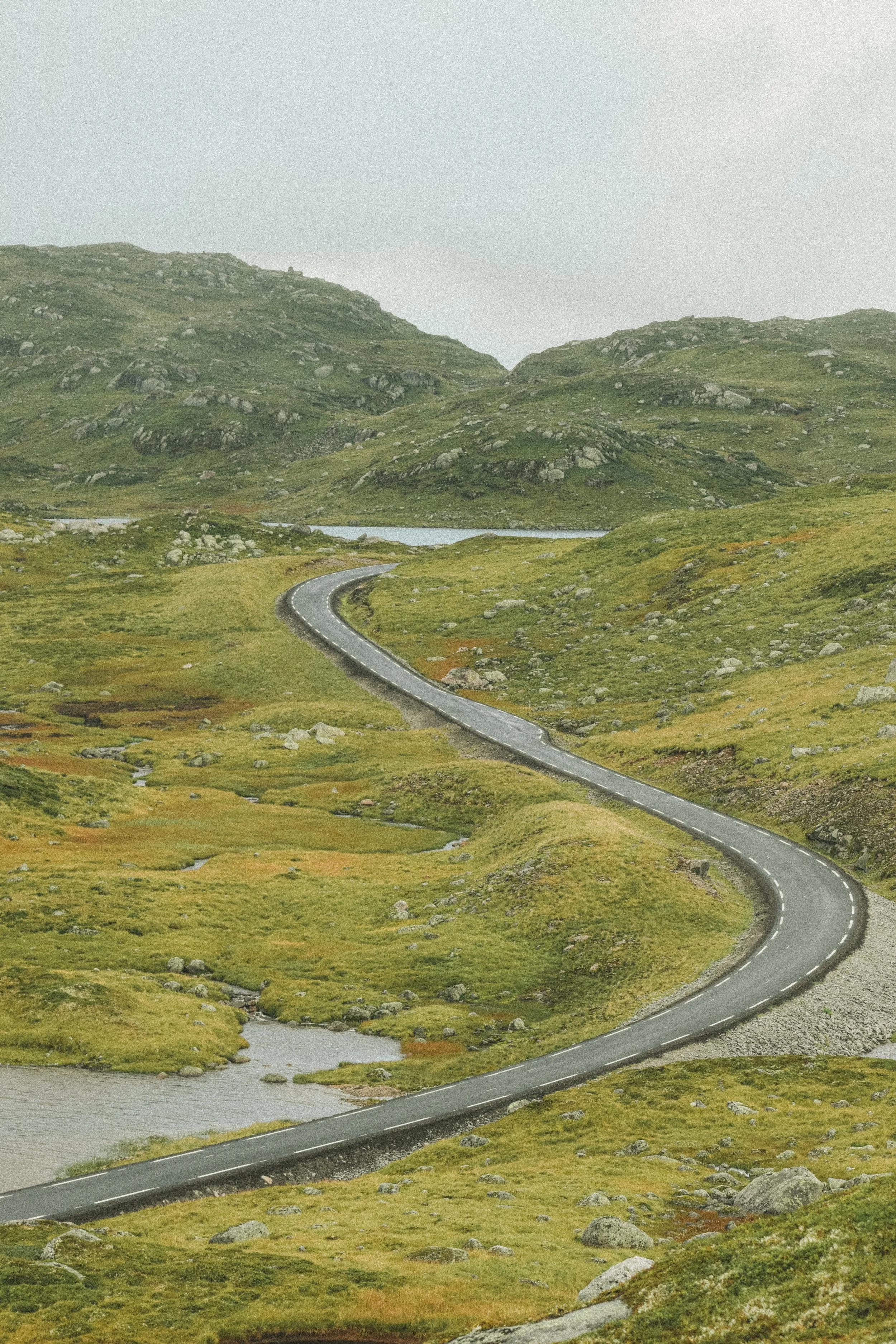 Paysage de montagnes avec une route sinueuse traversant des prairies verdoyantes, rivière et petit lac sous un ciel nuageux.