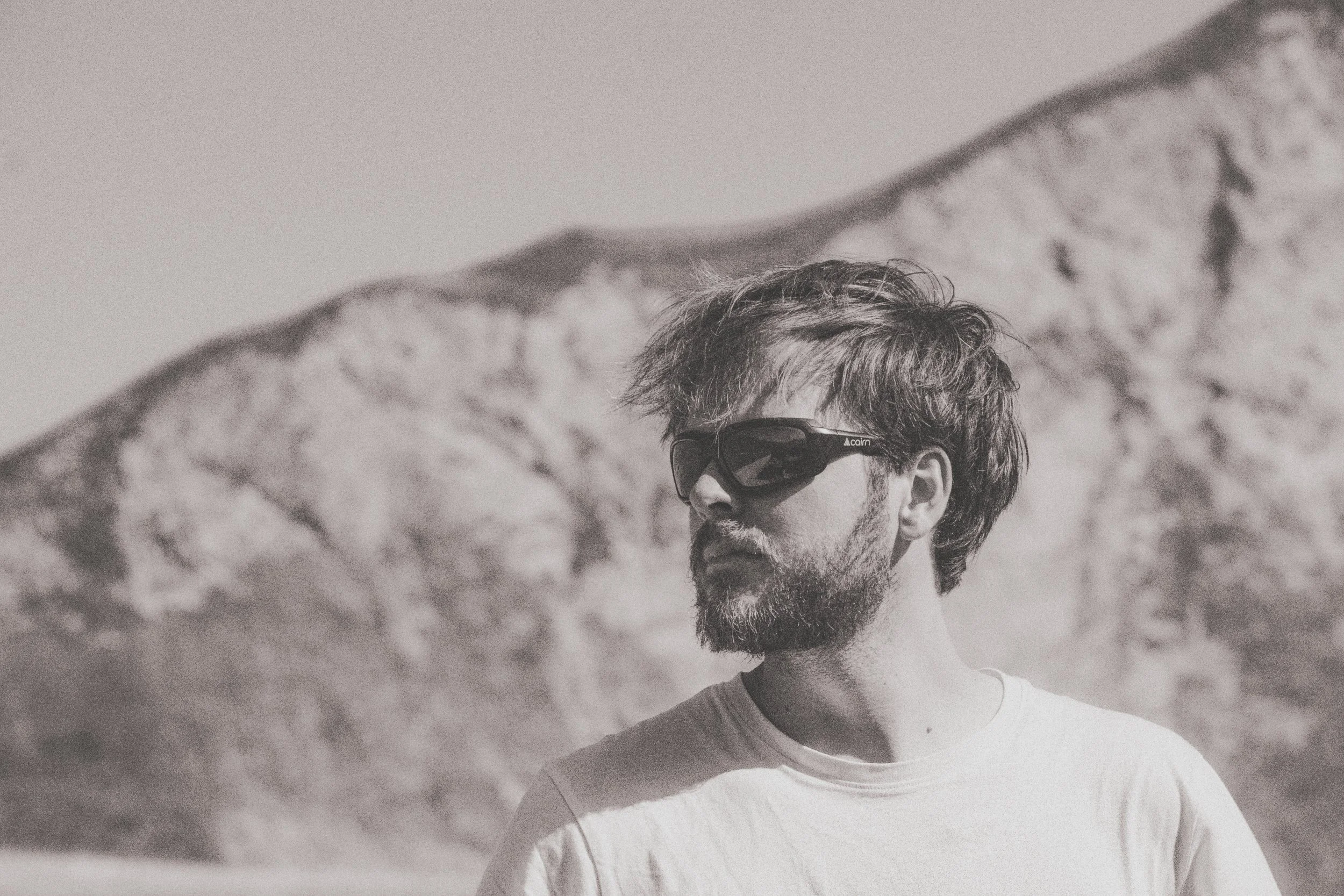 homme avec lunettes de soleil, barbe et moustache, regardant vers la droite, paysage montagneux en arrière-plan