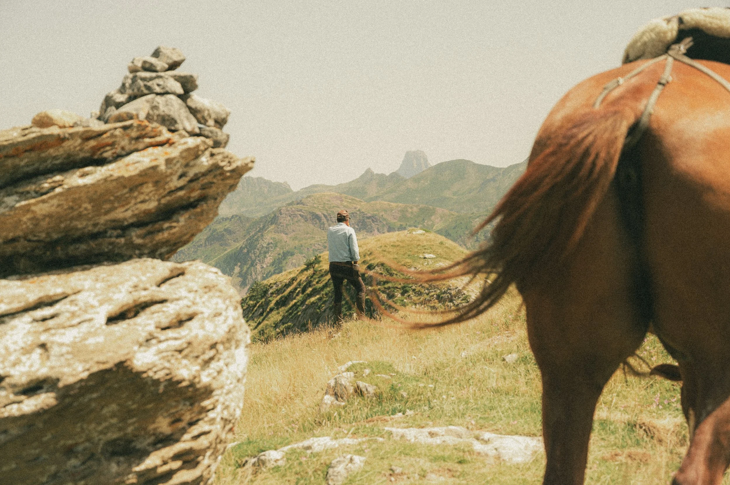 Un homme marche dans une vallée de montagne, vue partielle du dos d'un cheval en premier plan à droite, avec des rochers en premier plan à gauche.