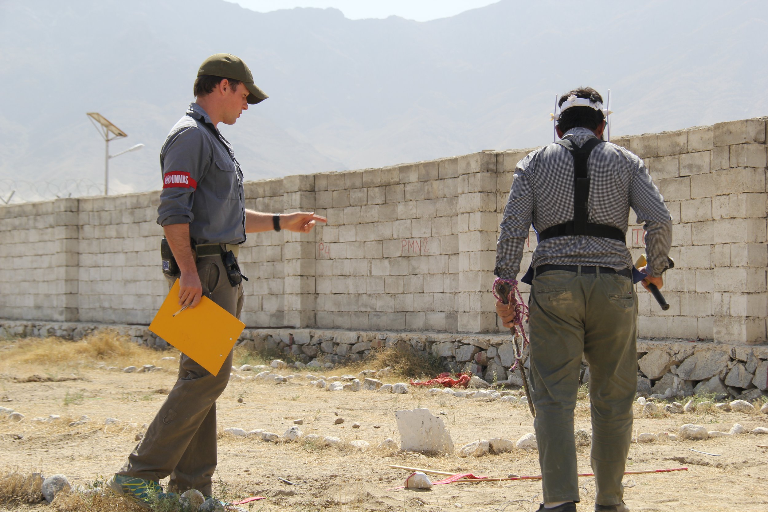  Two individuals, equipped with metal detectors, carefully scan a sandy area for mines against the backdrop of a high wall, exemplifying the hands-on aspect of Artios Global’s capacity development in mine action. 