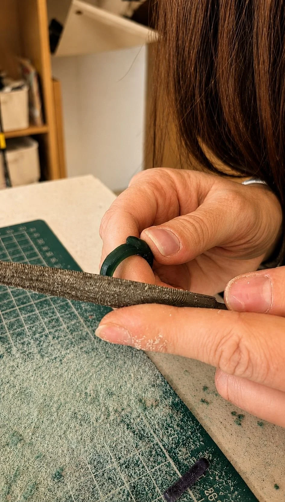Person working on a wax, using a file to shape and  smooth a wax carved ring, with a cutting mat and desk in the background.