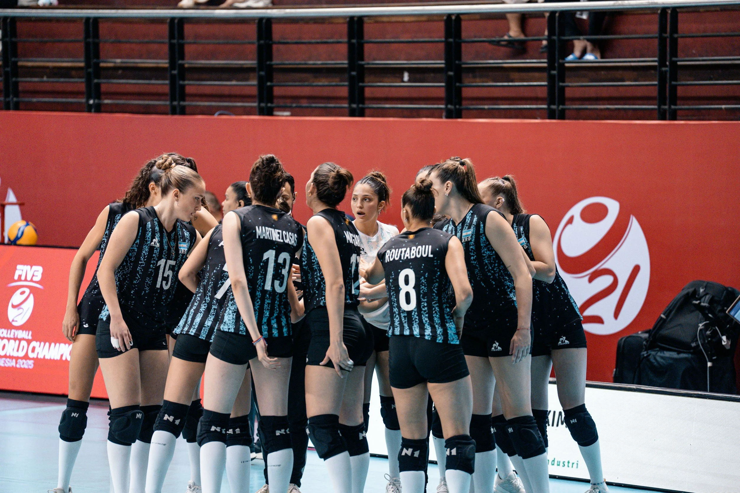 A women's volleyball team huddling up during a timeout. They are wearing dark and light blue uniforms, dark blue kneepads and white socks.