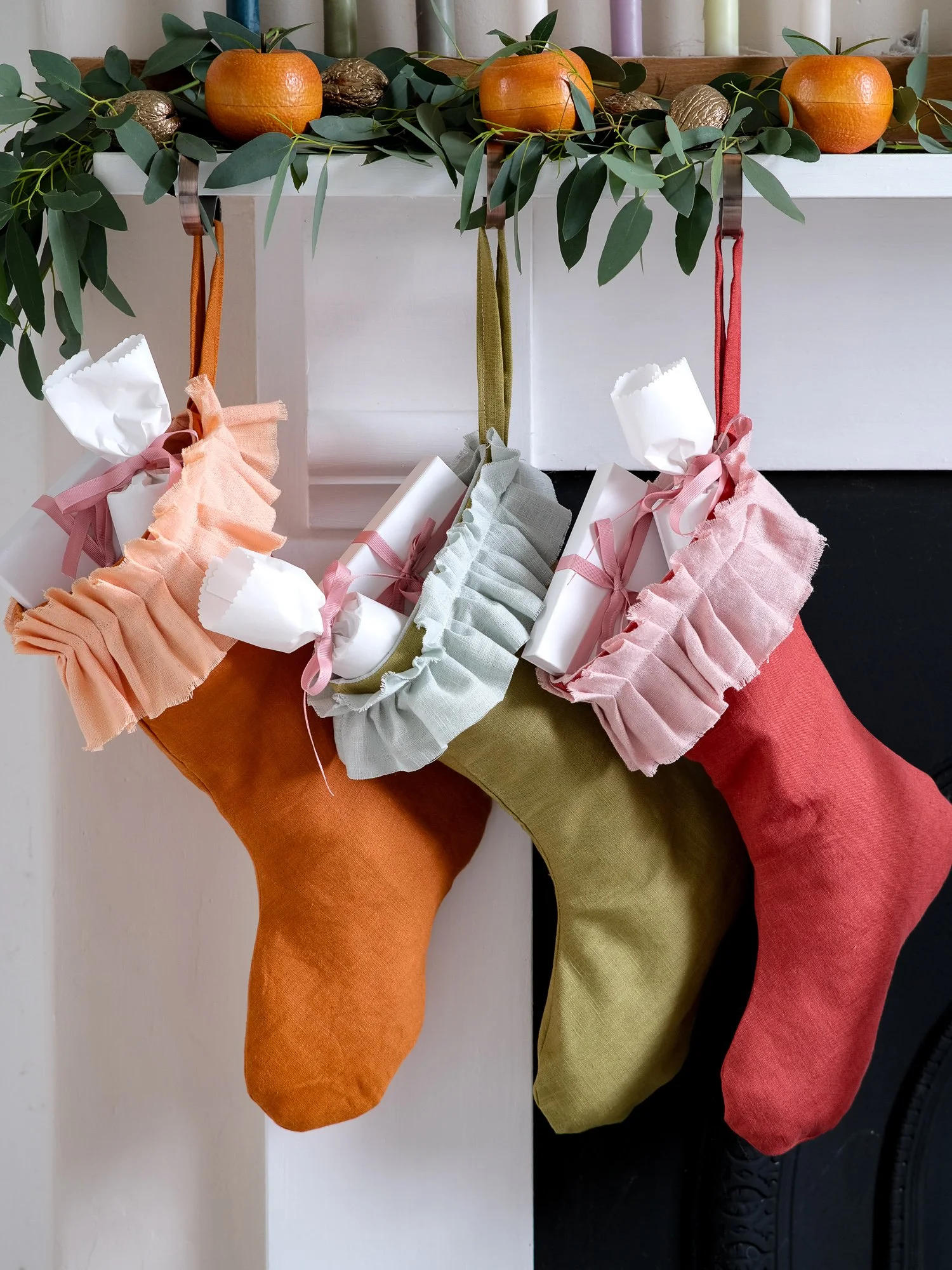 handmade linen Christmas stockings in warm orange, olive green and red hang beneath a festive mantel decorated with eucalyptus, oranges and candles, each finished with a soft, gathered ruffle cuff.