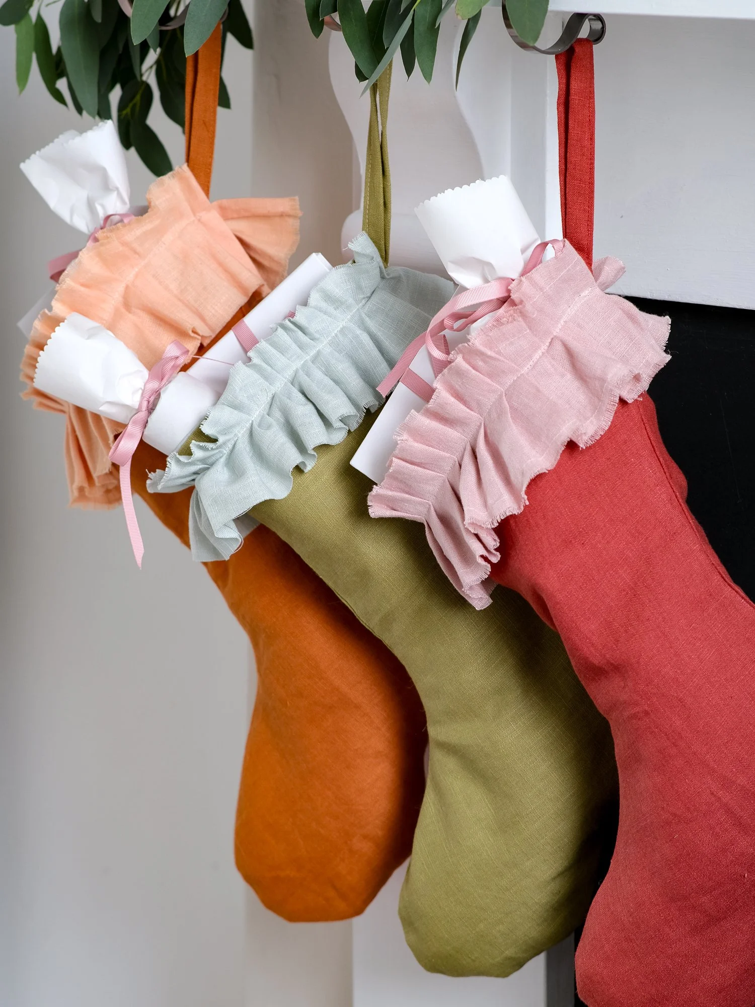 handmade linen Christmas stockings in warm orange, olive green and red hang beneath a festive mantel decorated with eucalyptus, oranges and candles, each finished with a soft, gathered ruffle cuff.