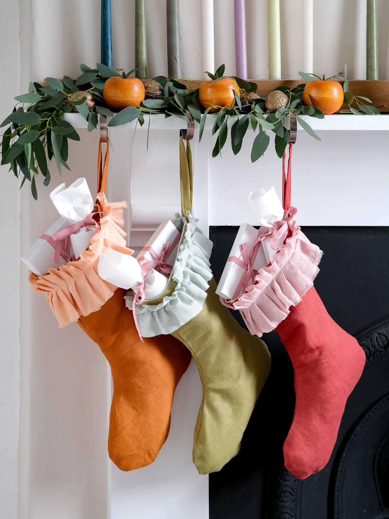 handmade linen Christmas stockings in warm orange, olive green and red hang beneath a festive mantel decorated with eucalyptus, oranges and candles, each finished with a soft, gathered ruffle cuff.