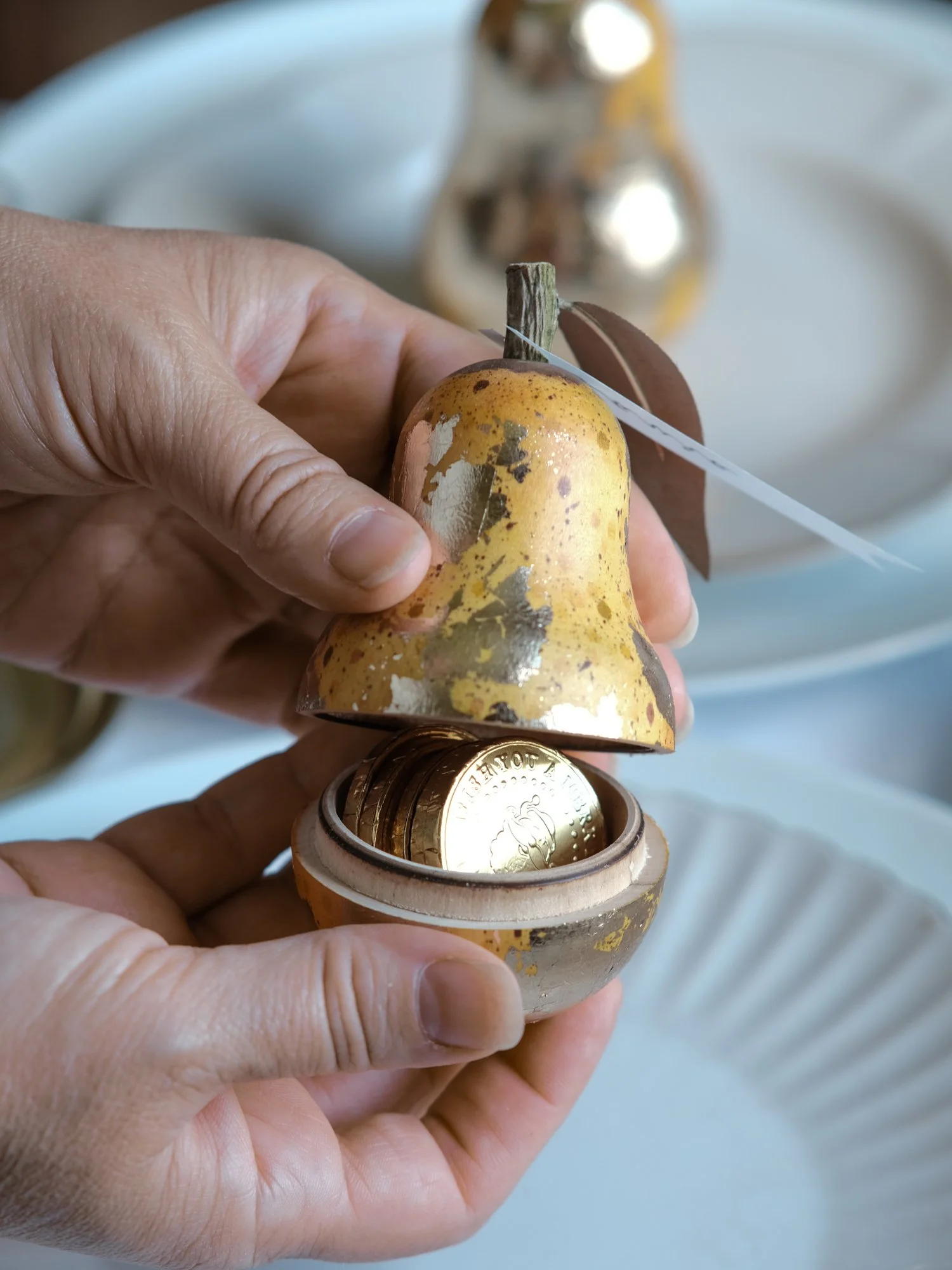 Gilded vintage-style Christmas pear ornament opened to reveal a small surprise inside, shown on a festive Christmas table setting with white plates and ribbon.