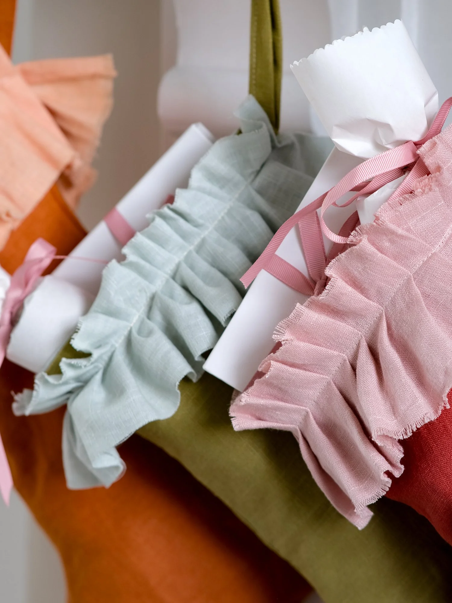 handmade linen Christmas stockings in warm orange, olive green and red hang beneath a festive mantel decorated with eucalyptus, oranges and candles, each finished with a soft, gathered ruffle cuff.