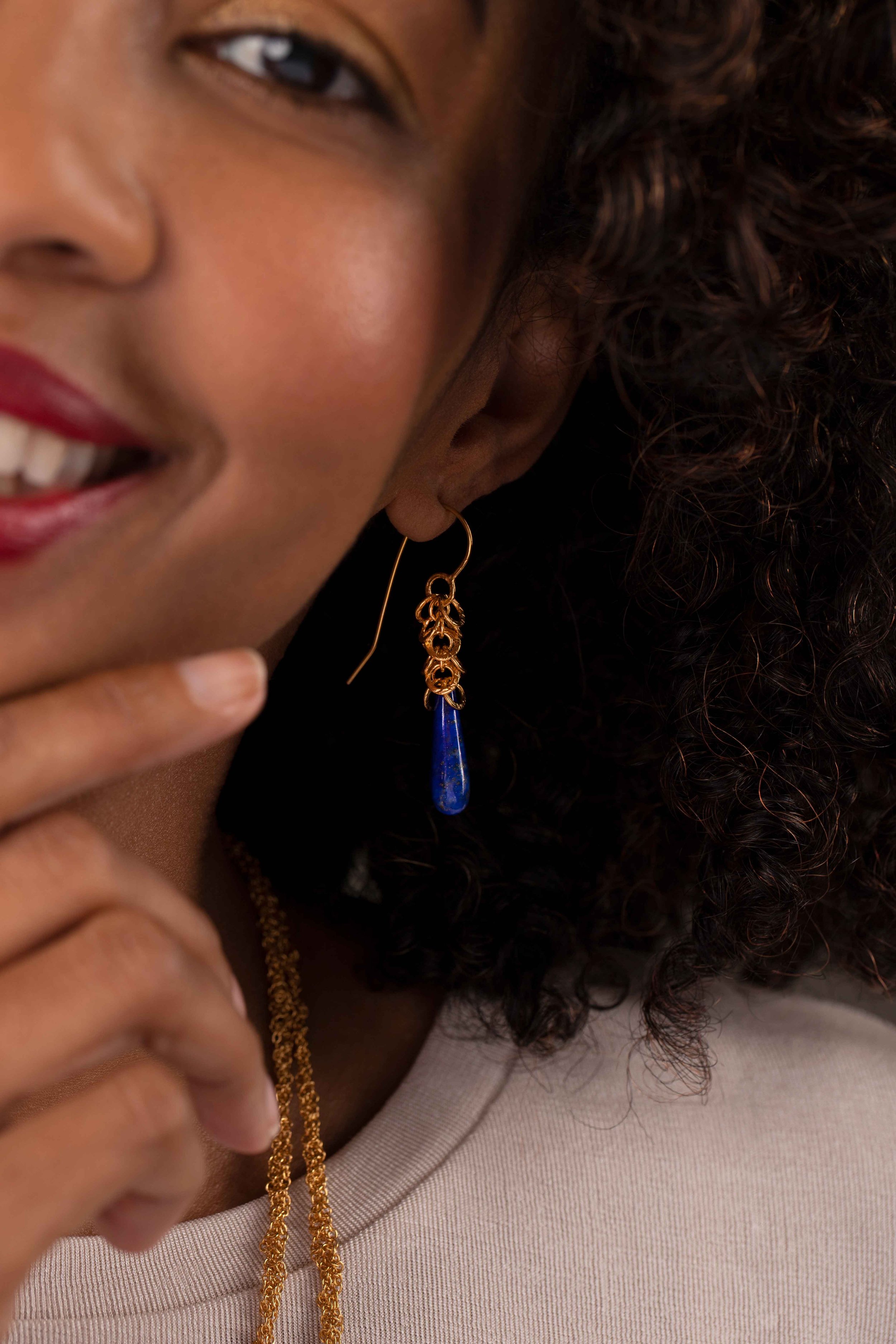 Close-up of a person wearing a gold earring with a blue teardrop pendant and a gold necklace, partially visible smile with red lipstick.