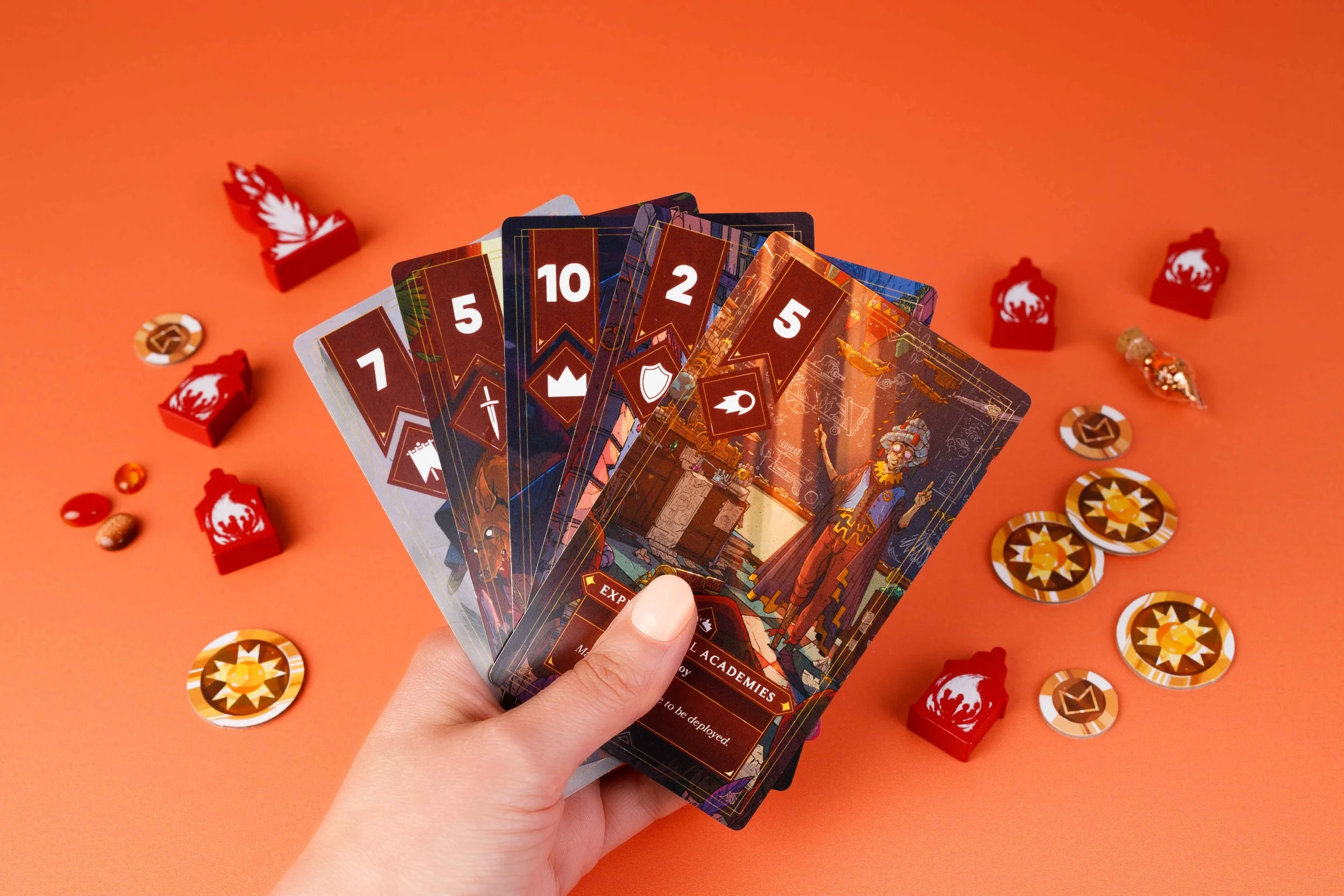 Hand holding fantasy-themed game cards, red tokens, and coins on an orange background.