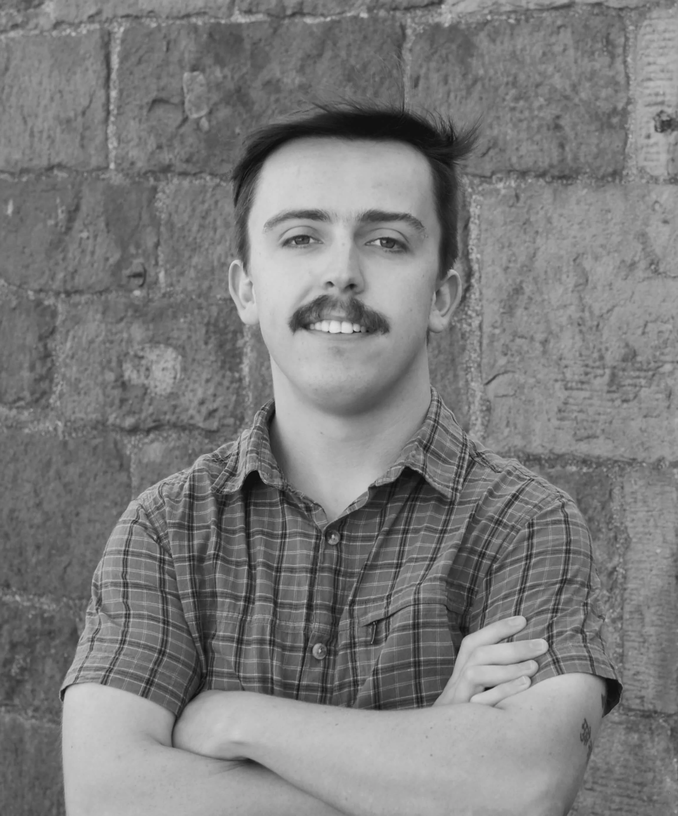Black and white portrait of a young man with a mustache, crossed arms, in front of a stone wall, wearing a plaid shirt.