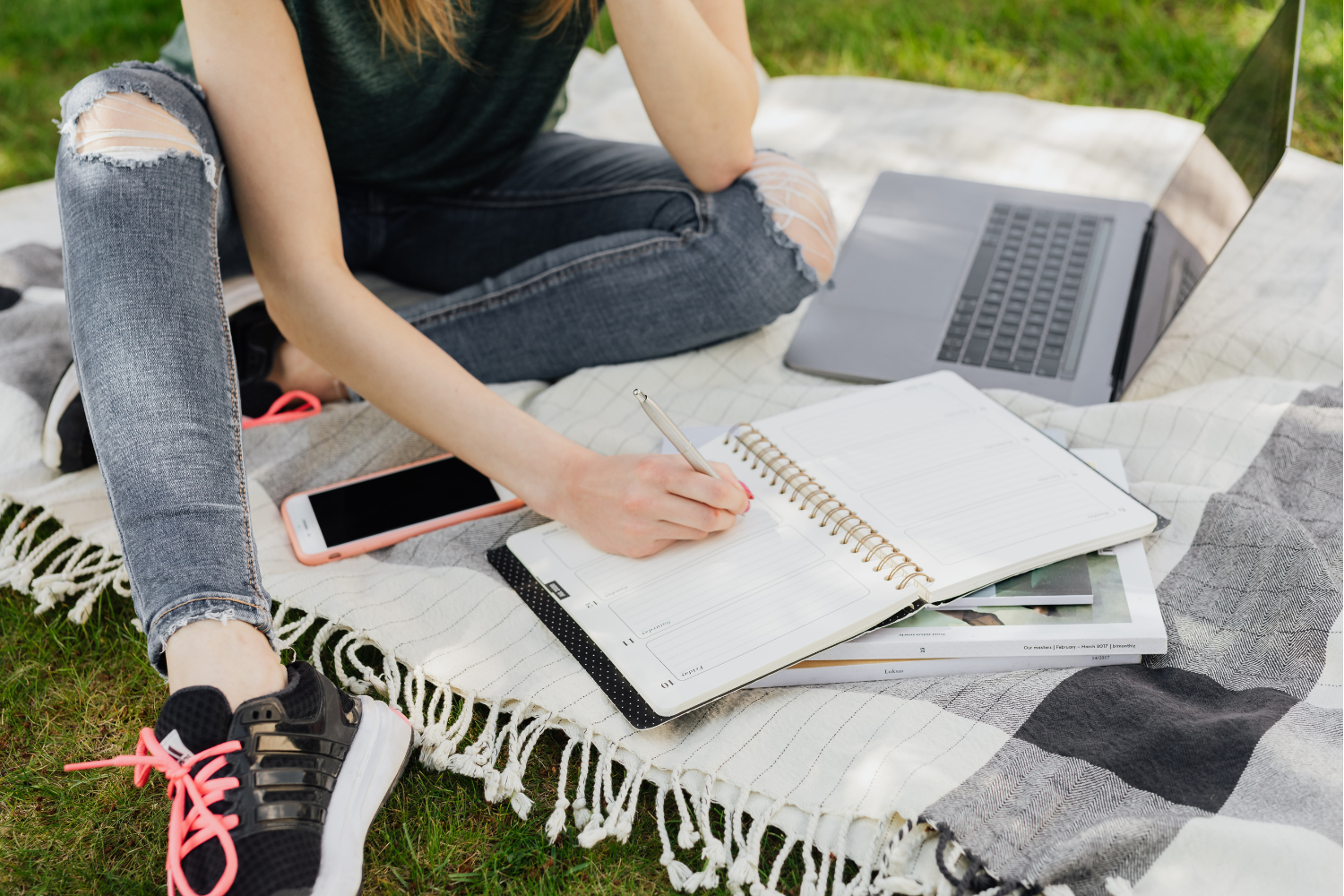 A teenager sitting on a blanket outdoors writing in a planner with a laptop nearby, representing summer goal setting and executive functioning skill building outside of the classroom.