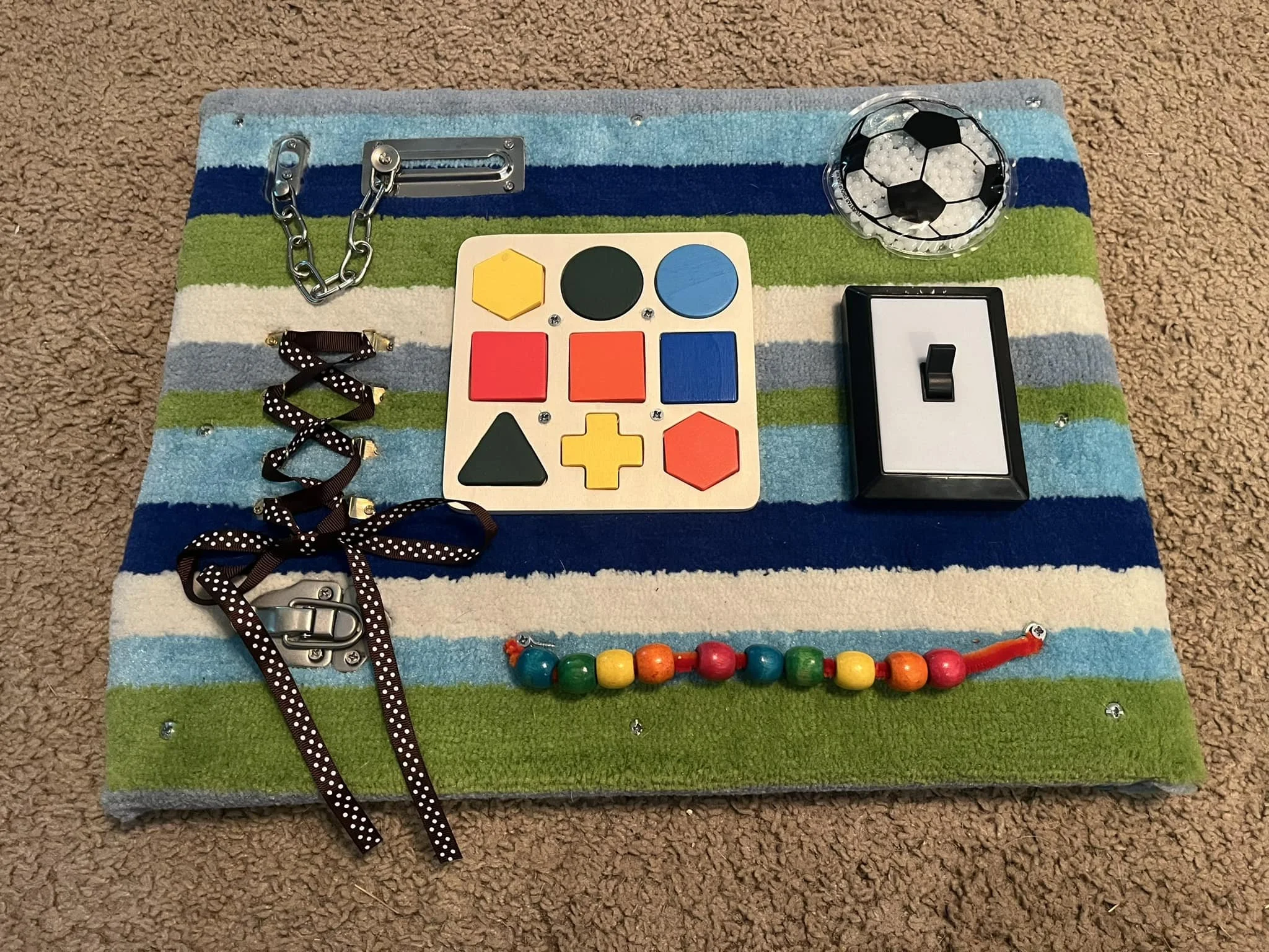 A colorful sensory and development activity board on a striped rug with various objects, including a black and white soccer ball, a mini black and white spiral, a switch, a wooden shape sorter with circle, square, and triangle shapes, a beaded string, and a chain with a buckle.