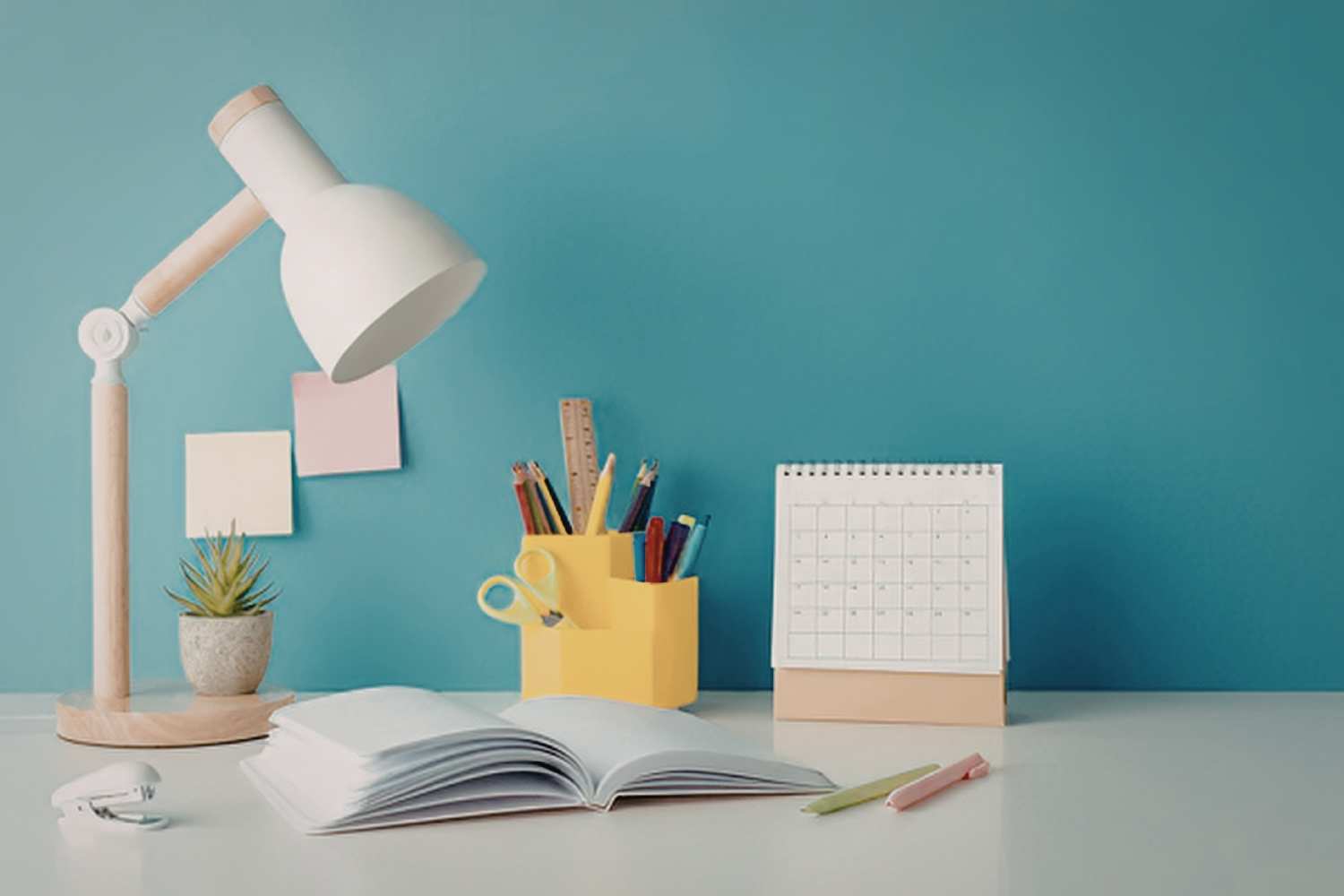 A bright, organized desk with an open notebook, calendar, colorful supplies, and a lamp against a teal wall — representing structure, planning, and organization skills supported through executive functioning coaching.