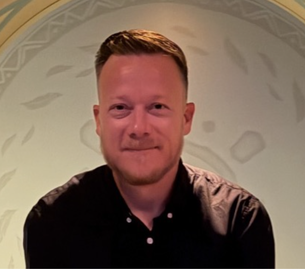 Close-up of a smiling man with short brown hair, wearing a black collared shirt, in front of a decorative background.