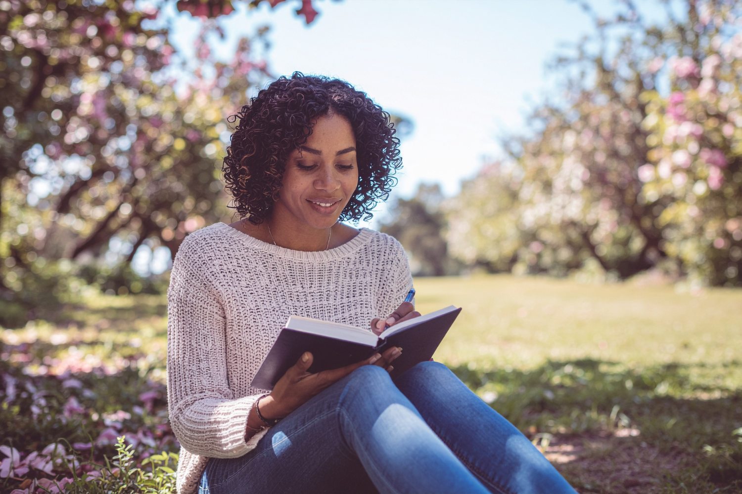 A woman sitting outdoors in a blooming park, smiling while writing in a journal — illustrating self-reflection and personal growth through executive functioning coaching.