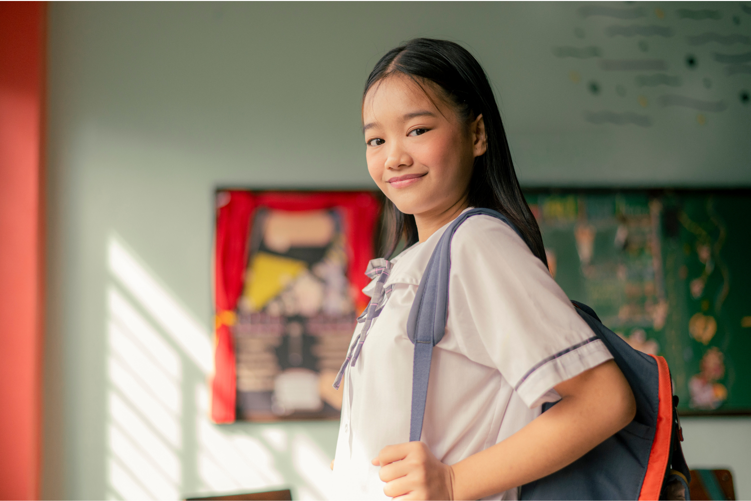 A confident young girl smiling with a backpack in a school classroom, representing a strong and prepared start to the school year following executive functioning coaching.