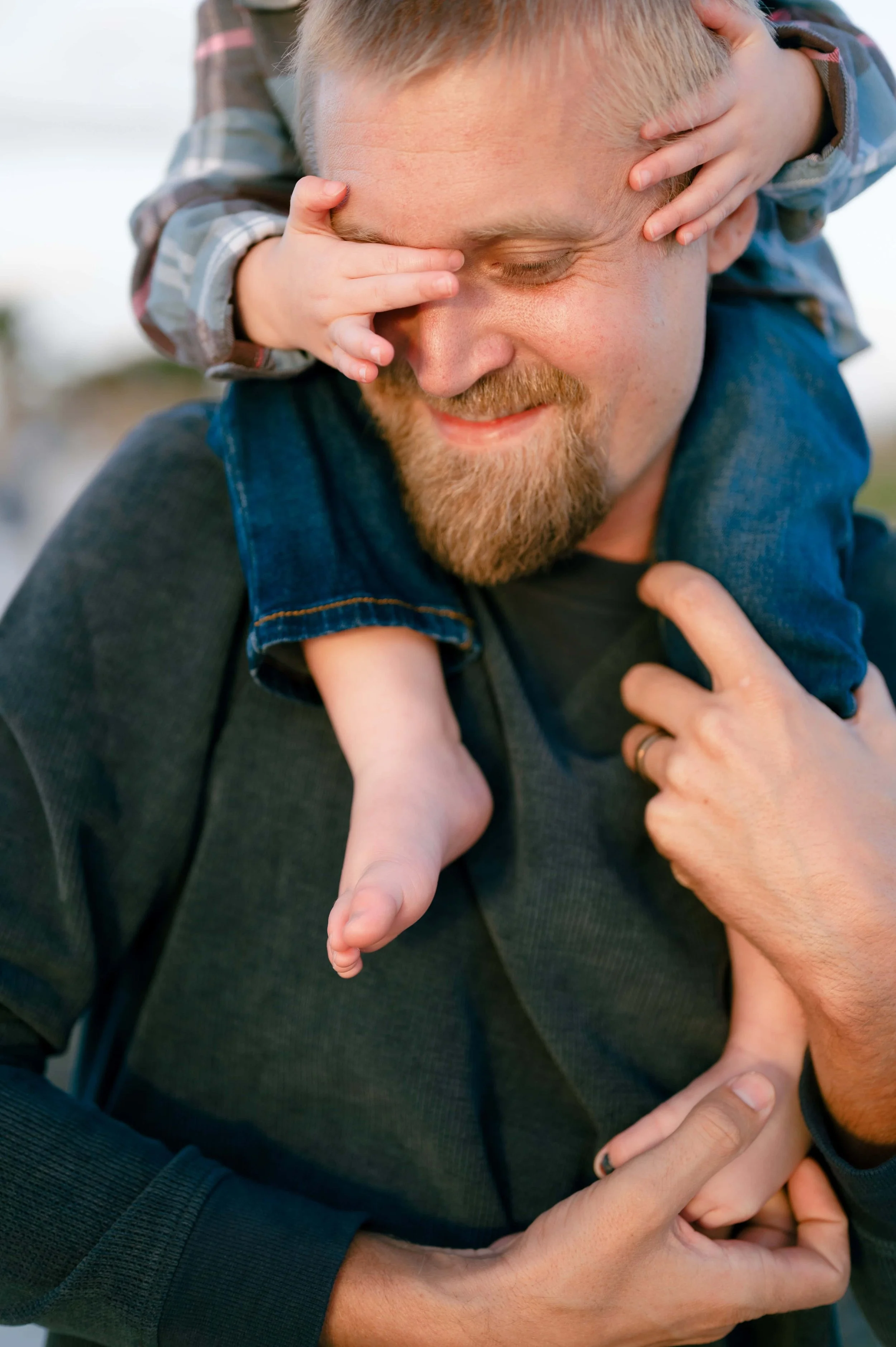 A man and a child enjoying a playful moment outdoors, with the child sitting on the man's shoulders and covering his eyes with hands, and the man smiling.
