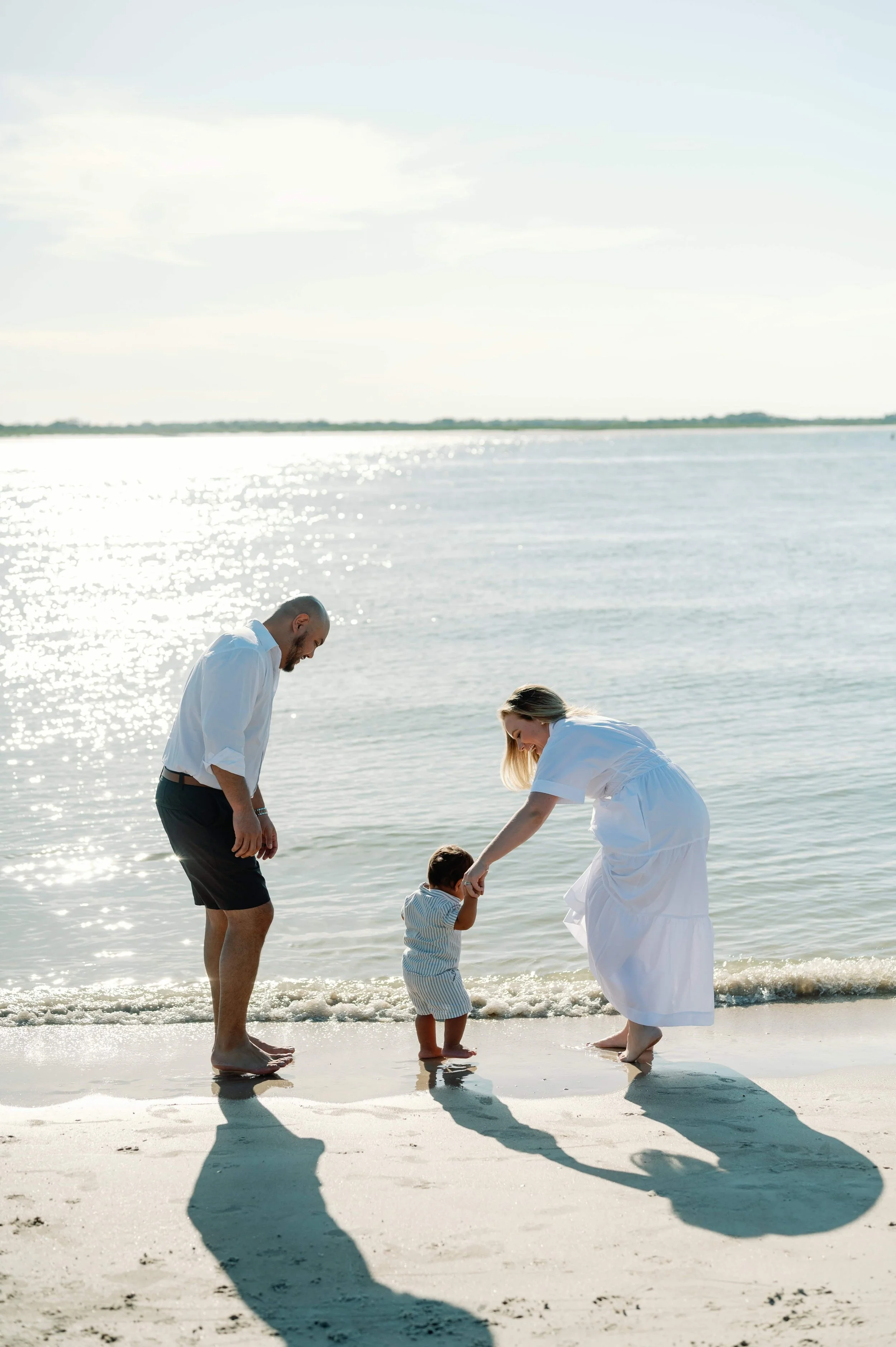 A family of three holding hands at the beach, with the woman and man leaning towards the child who is standing in the shallow water, all dressed in light clothing, under a sunny sky.