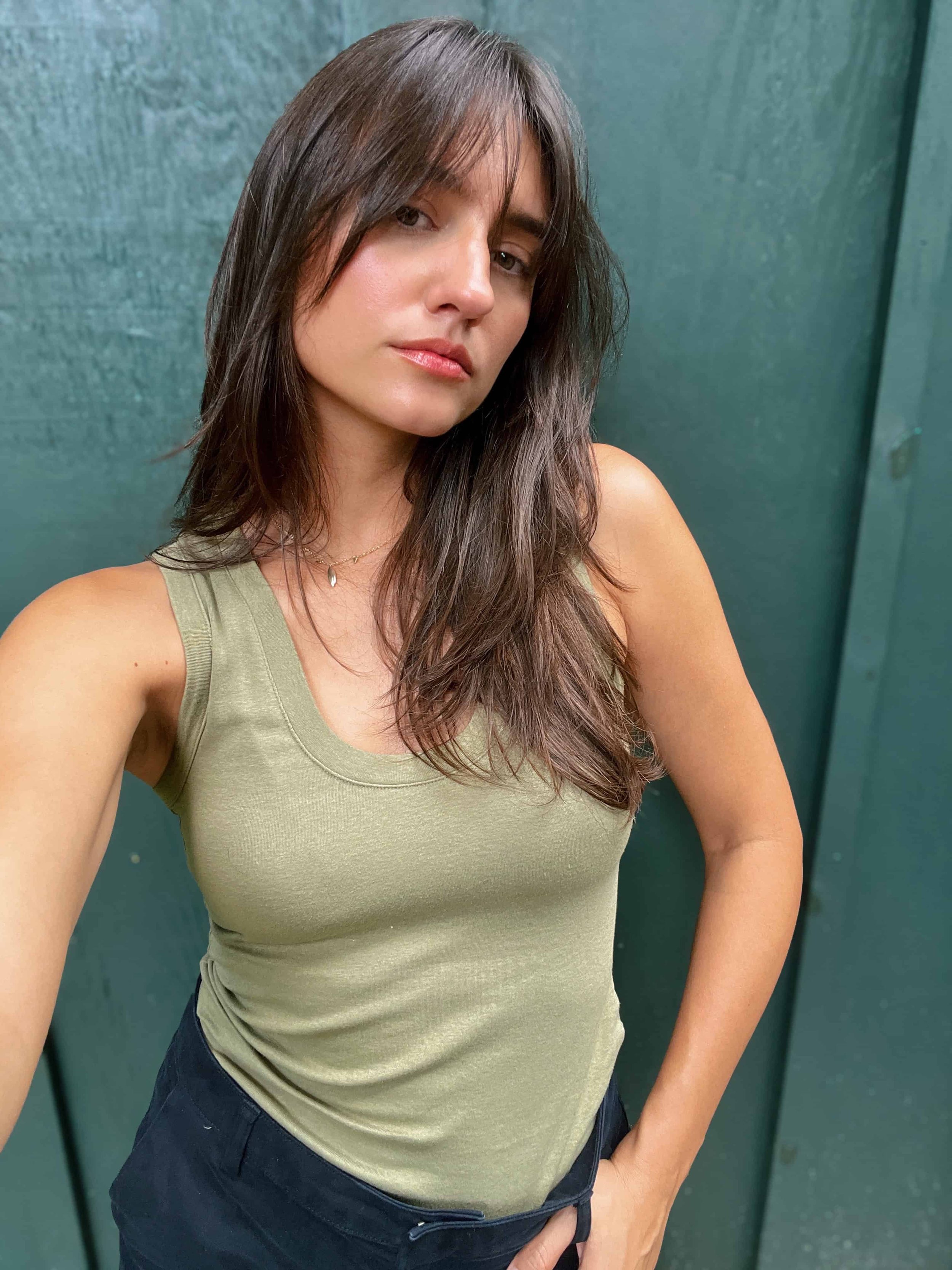 A woman with long brown hair wearing a green sleeveless top and black pants, standing against a green wall.
