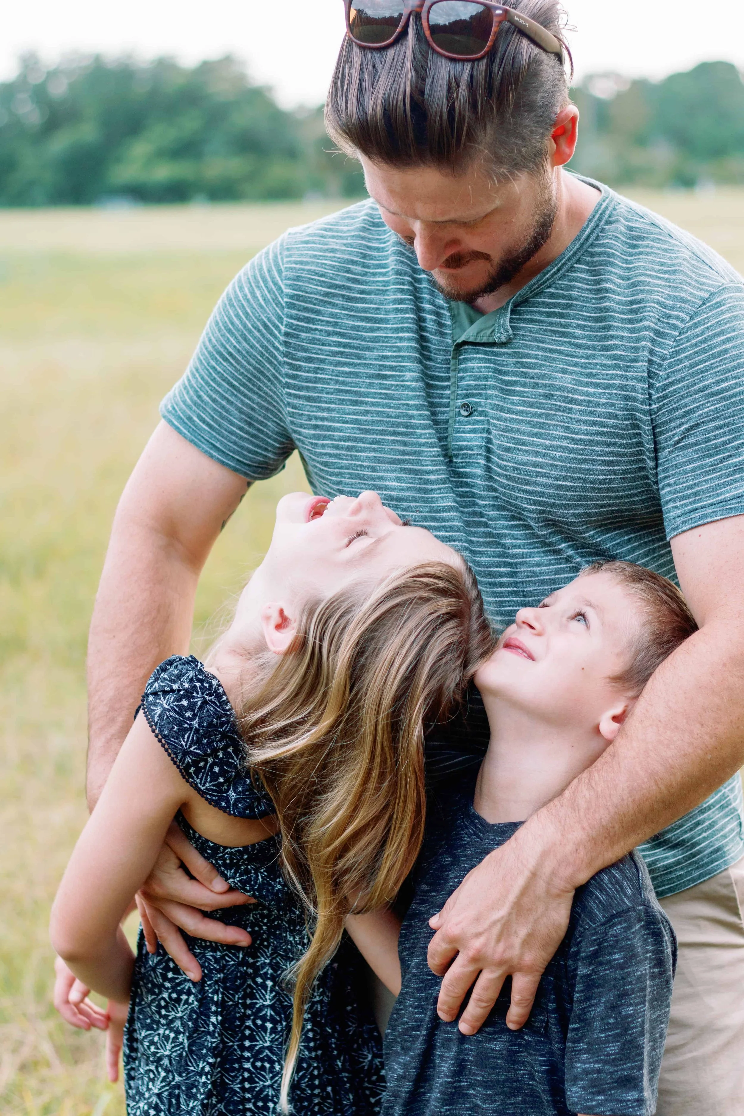 A man with two children, a girl and a boy, are outdoors in a field. The man is hugging the children, one on each side, and they are looking up at him happily. The setting appears to be a park or a meadow, with trees in the background.