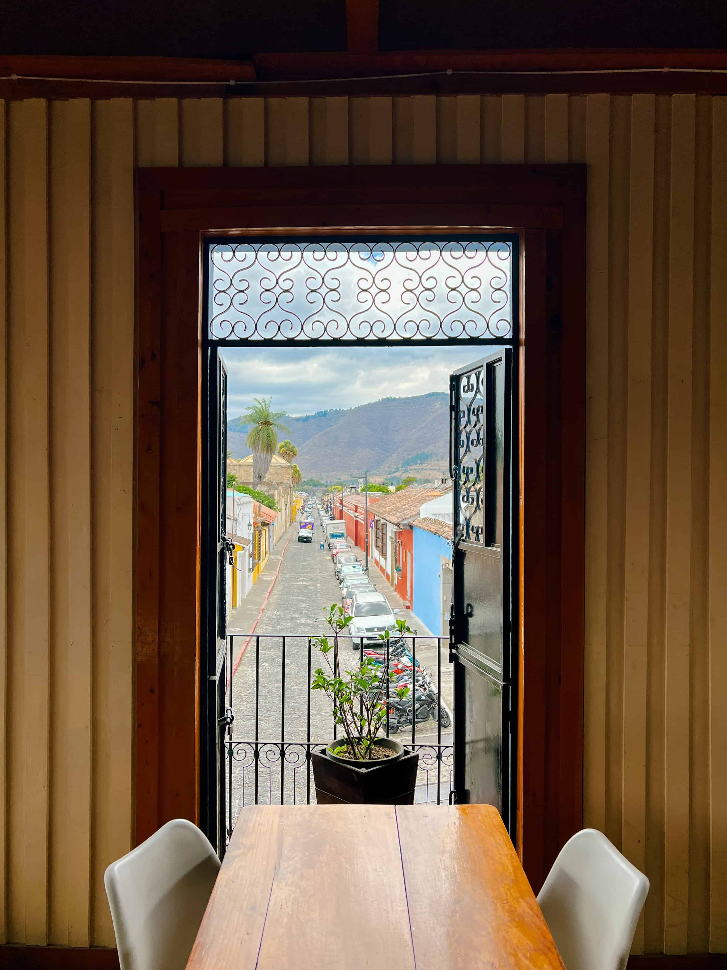 View from inside a room looking out a black wrought iron balcony door with open shutters onto a colorful street with parked cars and mountains in the background.