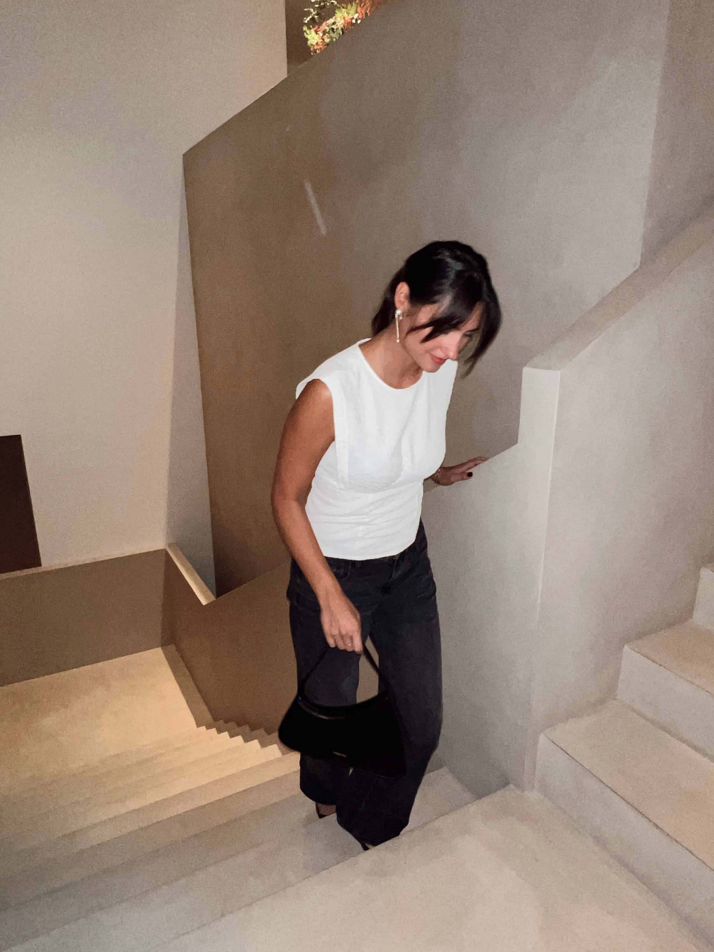 A woman with dark hair, wearing a white sleeveless top, black pants, and earrings, descending a beige staircase with a black purse in her hand.
