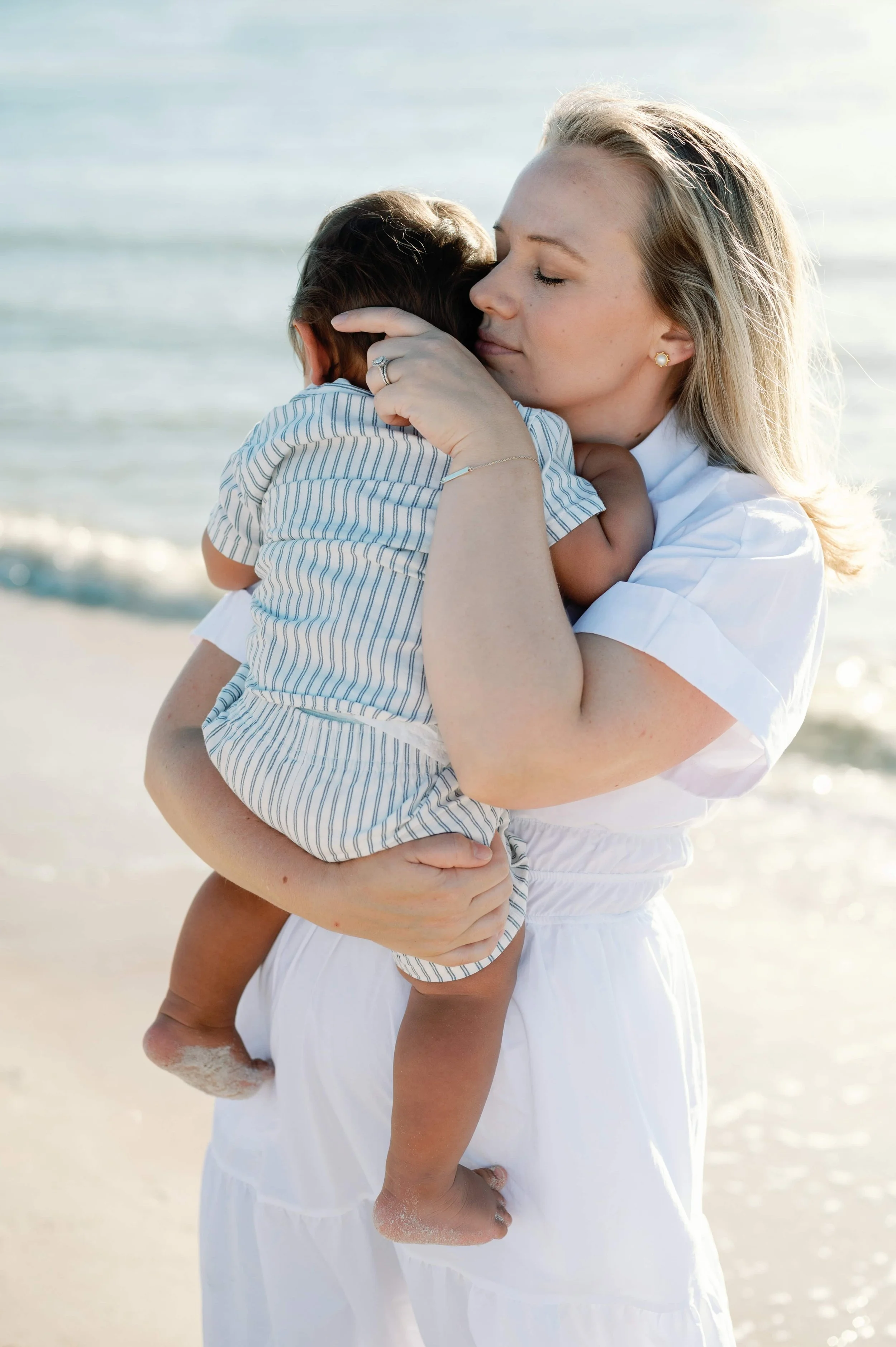 Woman holding a young child in a loving embrace at the beach with ocean in background.