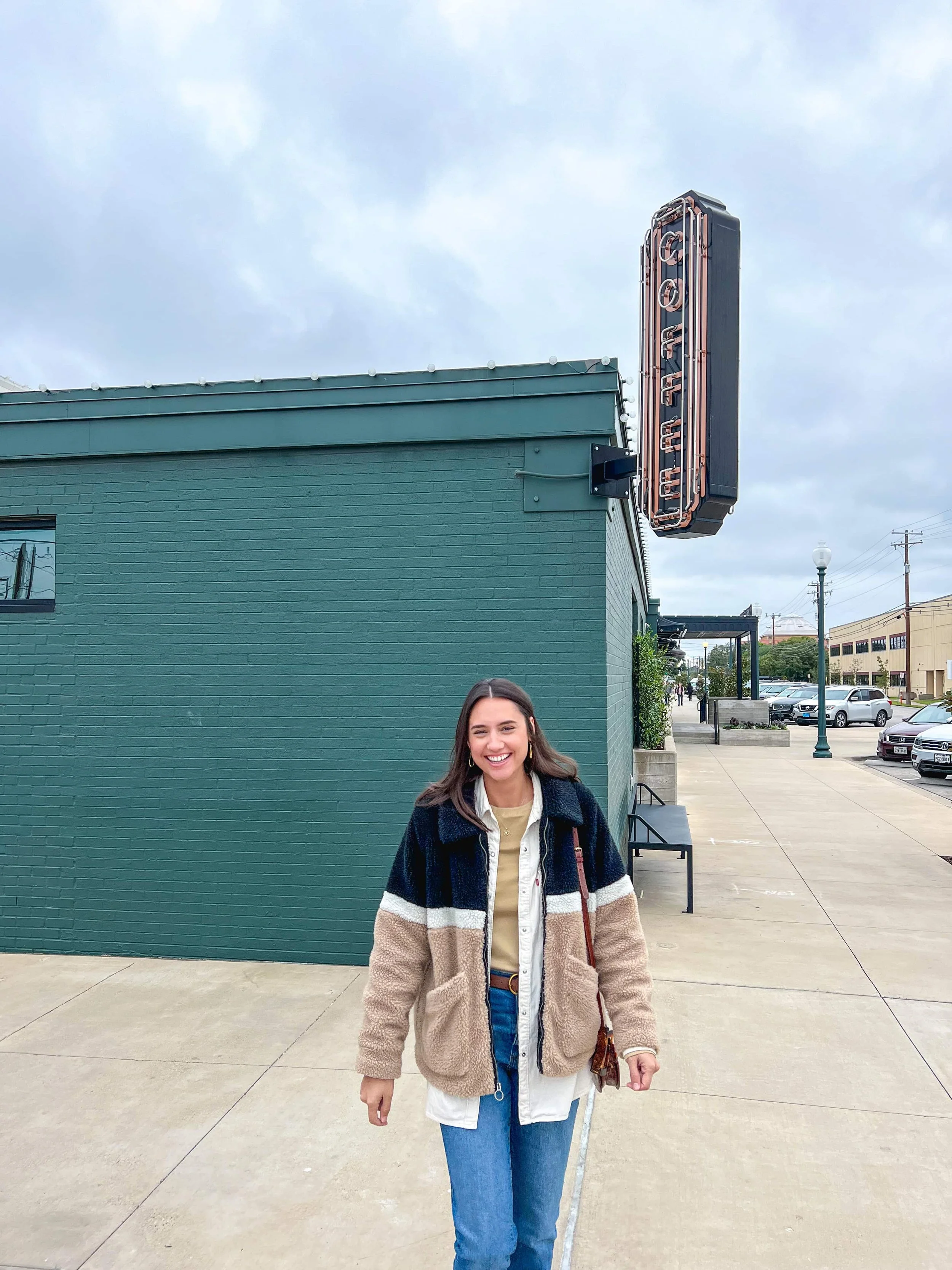 A woman with long brown hair is smiling and walking on a sidewalk in front of a dark green building with a neon sign that reads 'COFFEE.'