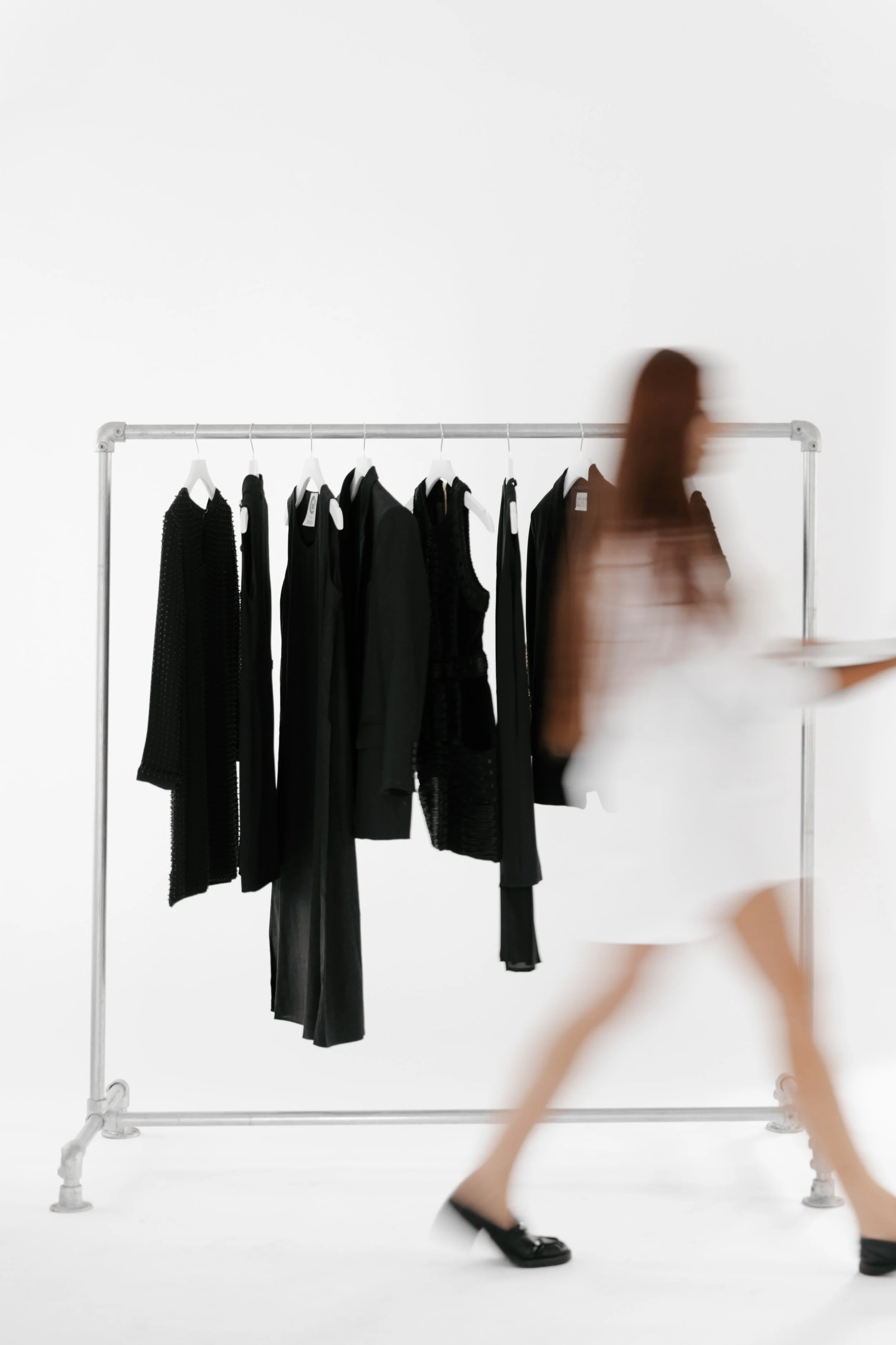 Clothing rack with black clothes in the background, white walls, and a fashion stylist walking by. DistriktF Park City Utah Photo Studio and Event Rental Space.