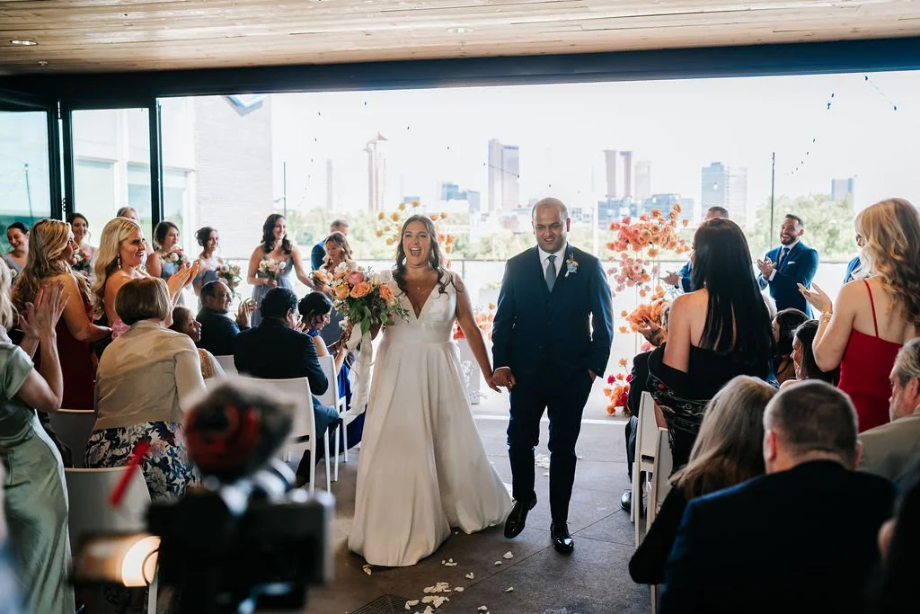 A bride and groom hold hands as they walk down their wedding ceremony aisle at The Fives Columbus. The city skyline is behind them and their guests are applauding.