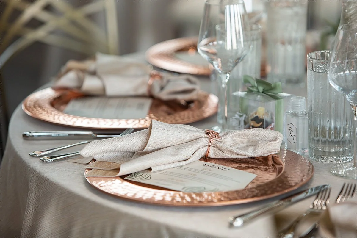 A wedding reception table is set with copper chargers and neutral napkins and linens.