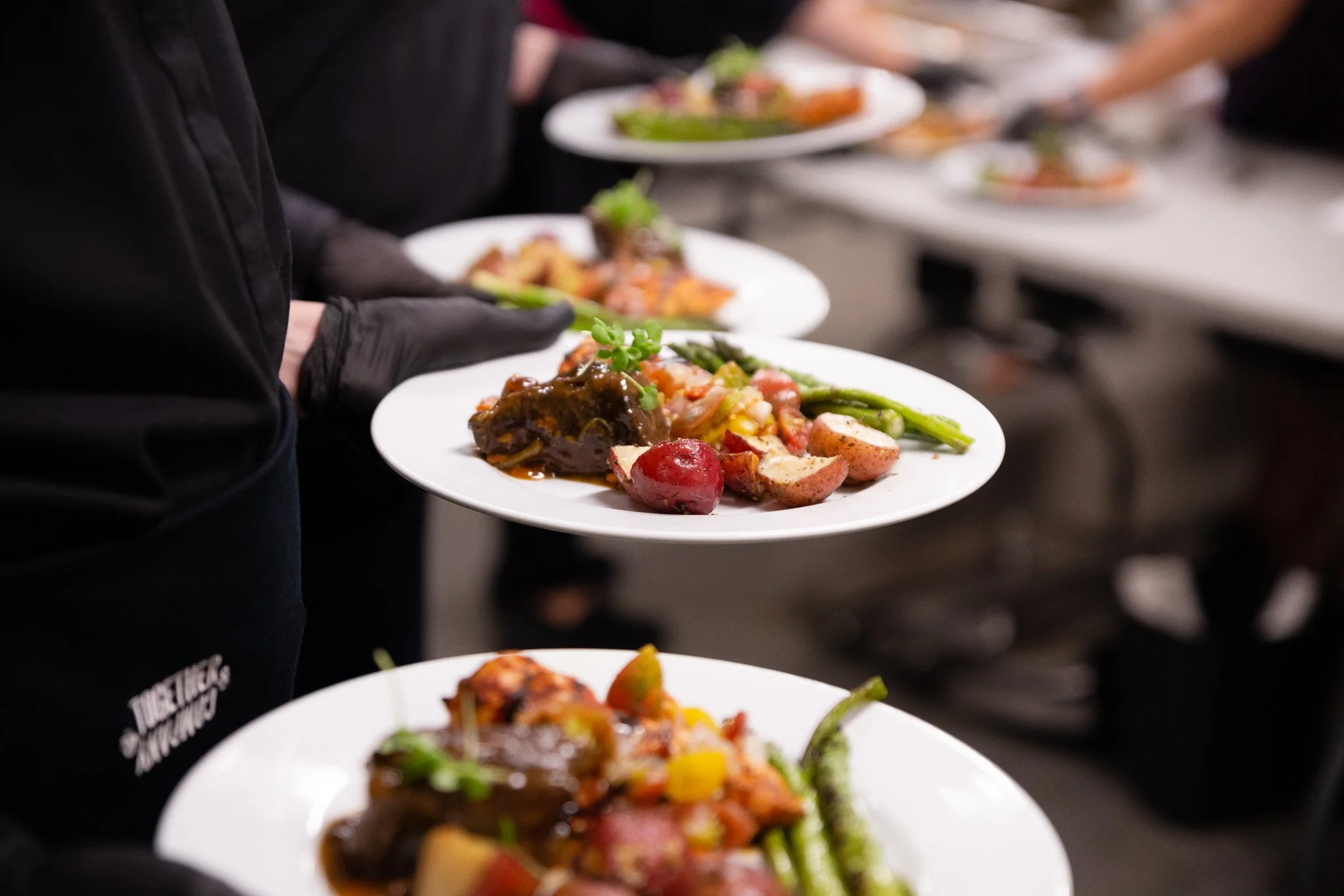 A line of servers hold plates of food at waist height, garnished and ready to serve. The focus is on one plate, and all the other plates align from the foreground to the background.
