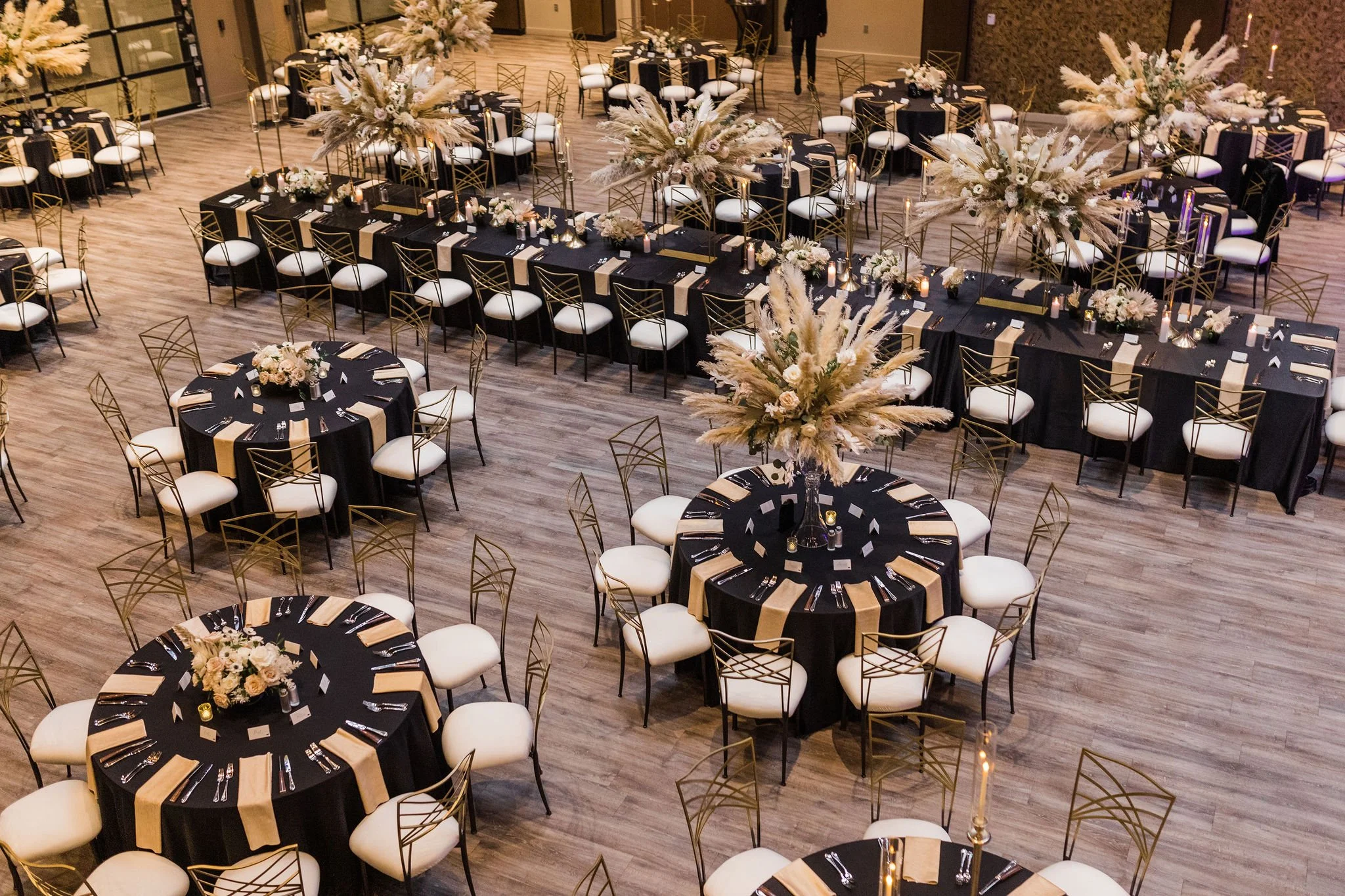 A wedding reception hall is set with round tables and one long banquet table. There are black linens on the tables and tall, neutral color centerpieces in the center of each table.