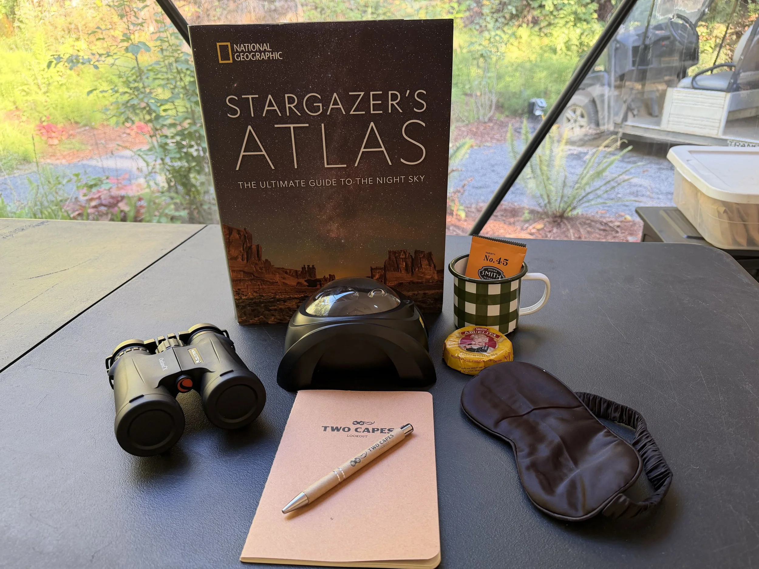 Items for stargazing laid out on a table include binoculars, a book, mug with tea, sleep mask, notebook with pen, hot cocoa, and a projector.