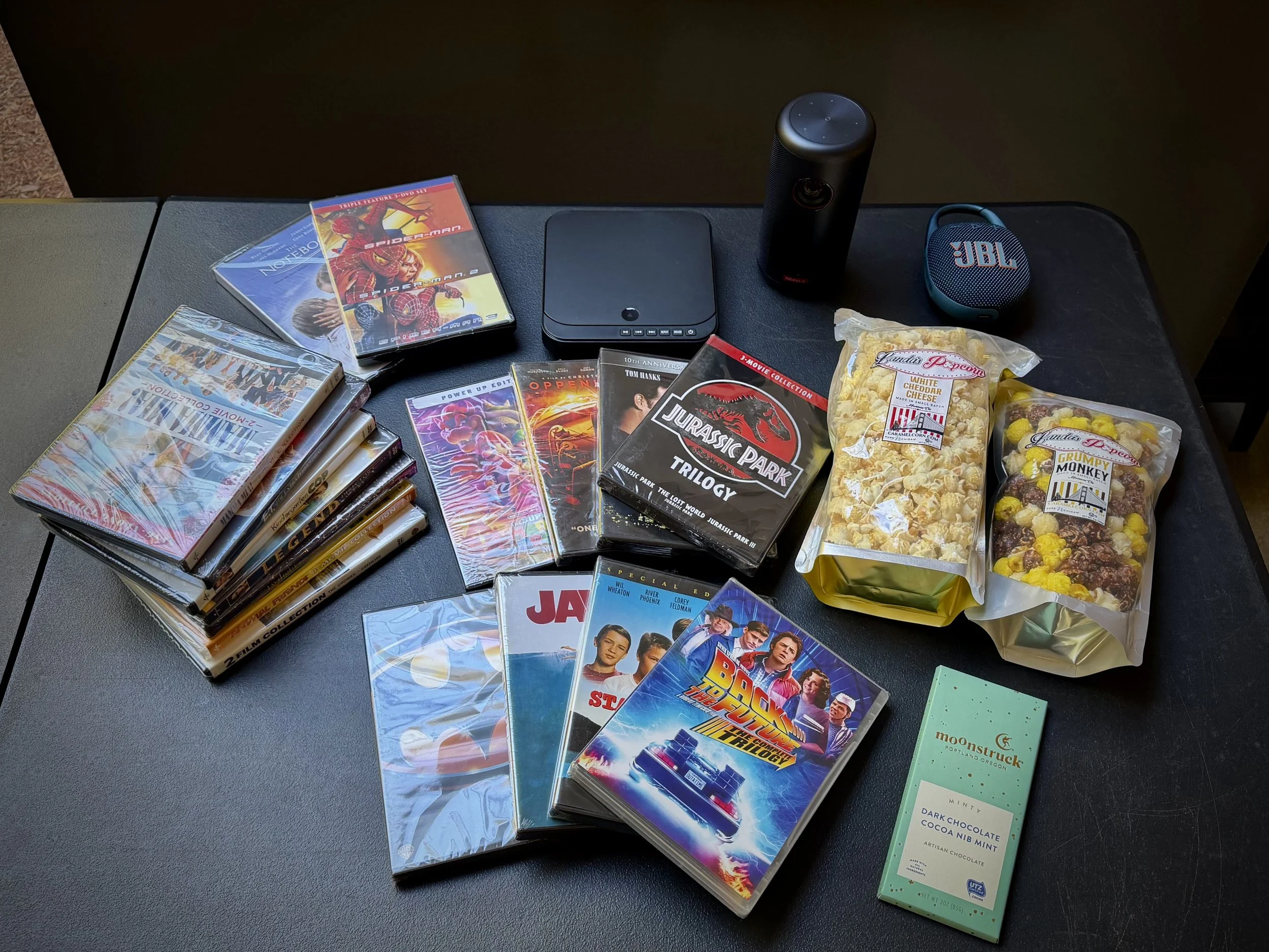 Items for a movie night are laid out on a table and include DVDs, two bags of gourmet popcorn, chocolate bar, blue ray player, speaker, and prejector.