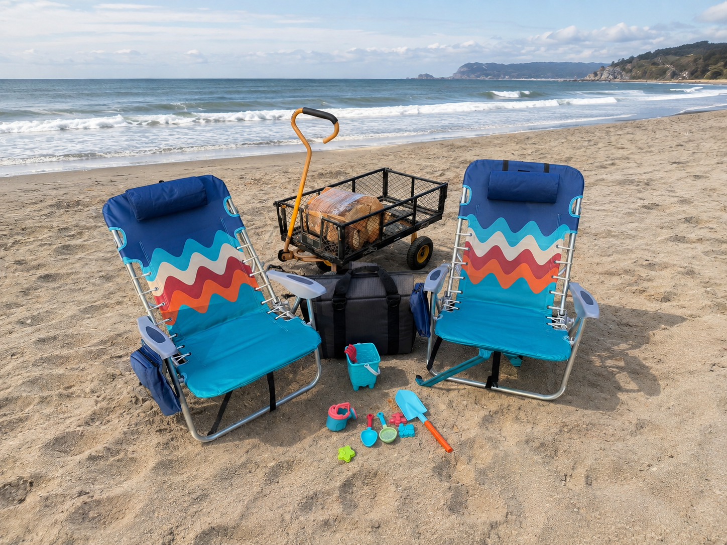 Two beach chairs are on the beach, with the ocean in the background. In between the chairs are sand toys, a soft-side cooler. Behind the chairs is a wagon with firewood.