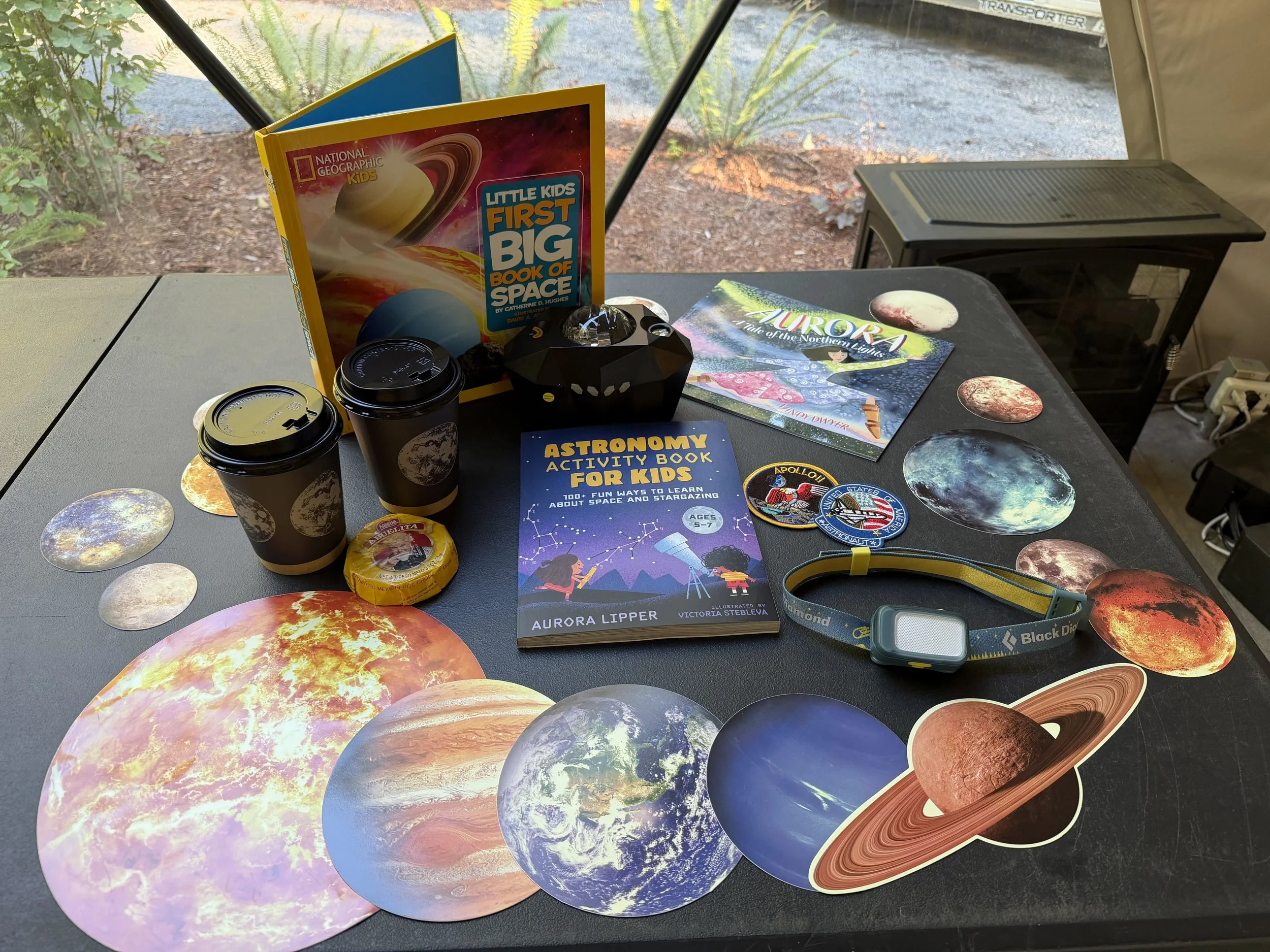 Space items are laid out on a table and include large stickers of the plants and sun, two books, a headlamp, patches, cups, cocoa, and a projector.