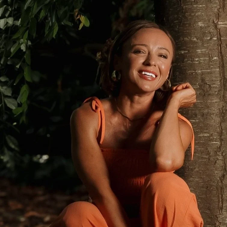 A woman in an orange dress smiling and leaning against a tree in a nighttime outdoor setting.