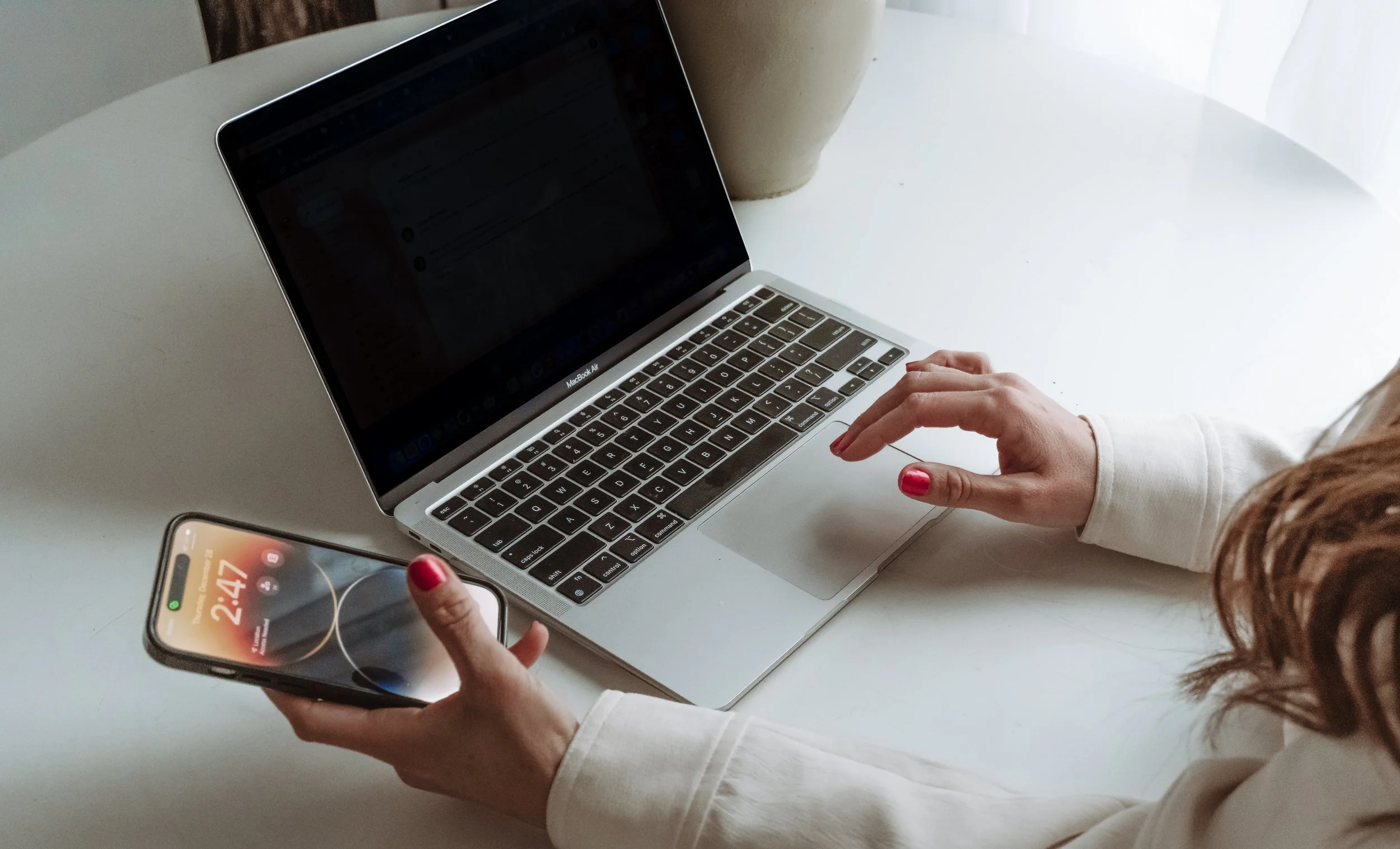 A person with red painted nails using a smartphone in their left hand and a silver MacBook Air on a white table.