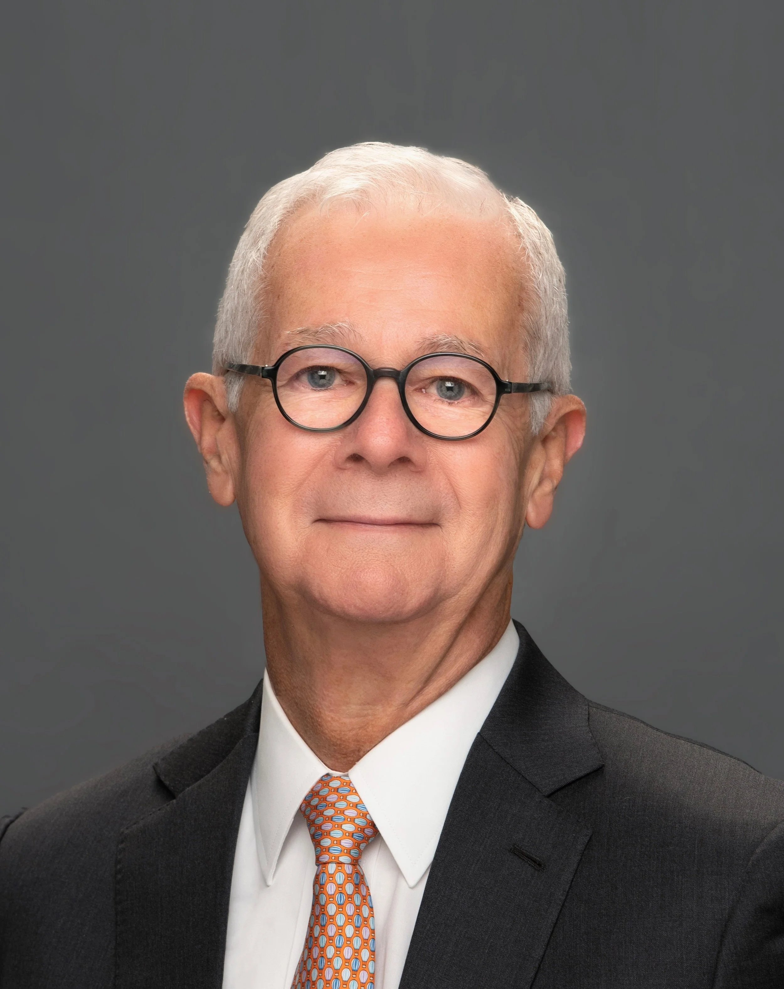 A professional headshot of an older man wearing glasses, a black suit, white shirt, and an orange patterned tie, against a gray background.