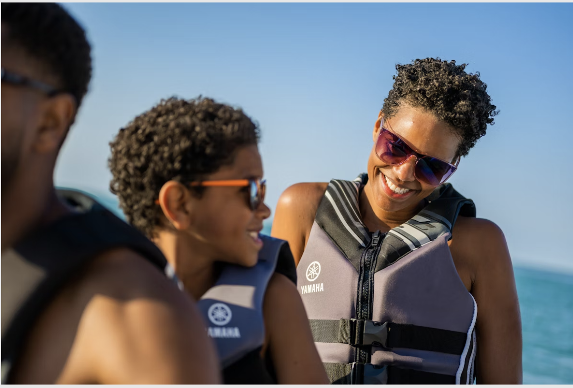 Three women in life jackets smiling and chatting outdoors with a blue sky and water in the background.