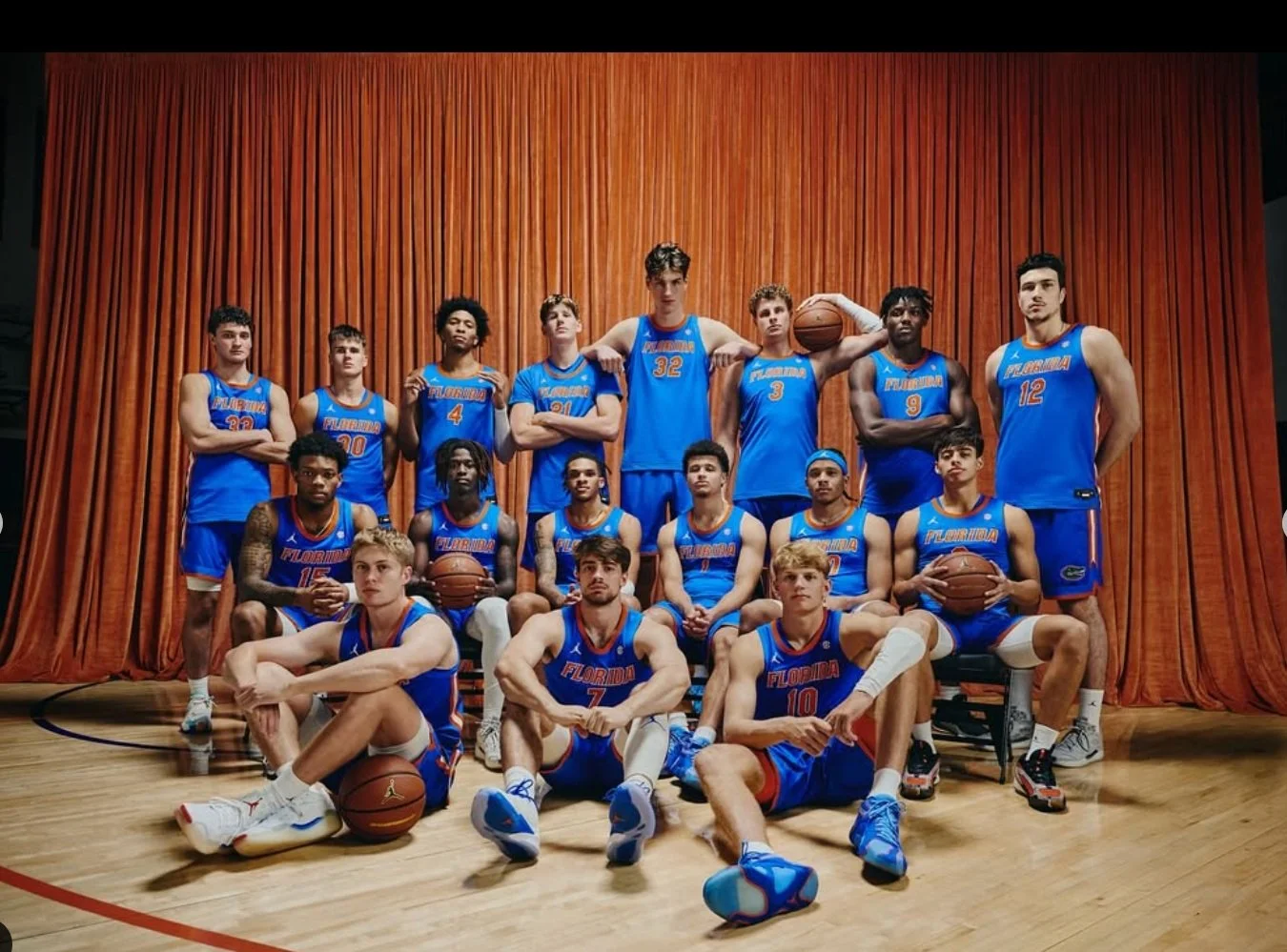 A basketball team in blue uniforms posing on a court with an orange curtain background. Eleven players are standing and five are seated or kneeling, with some holding basketballs.
